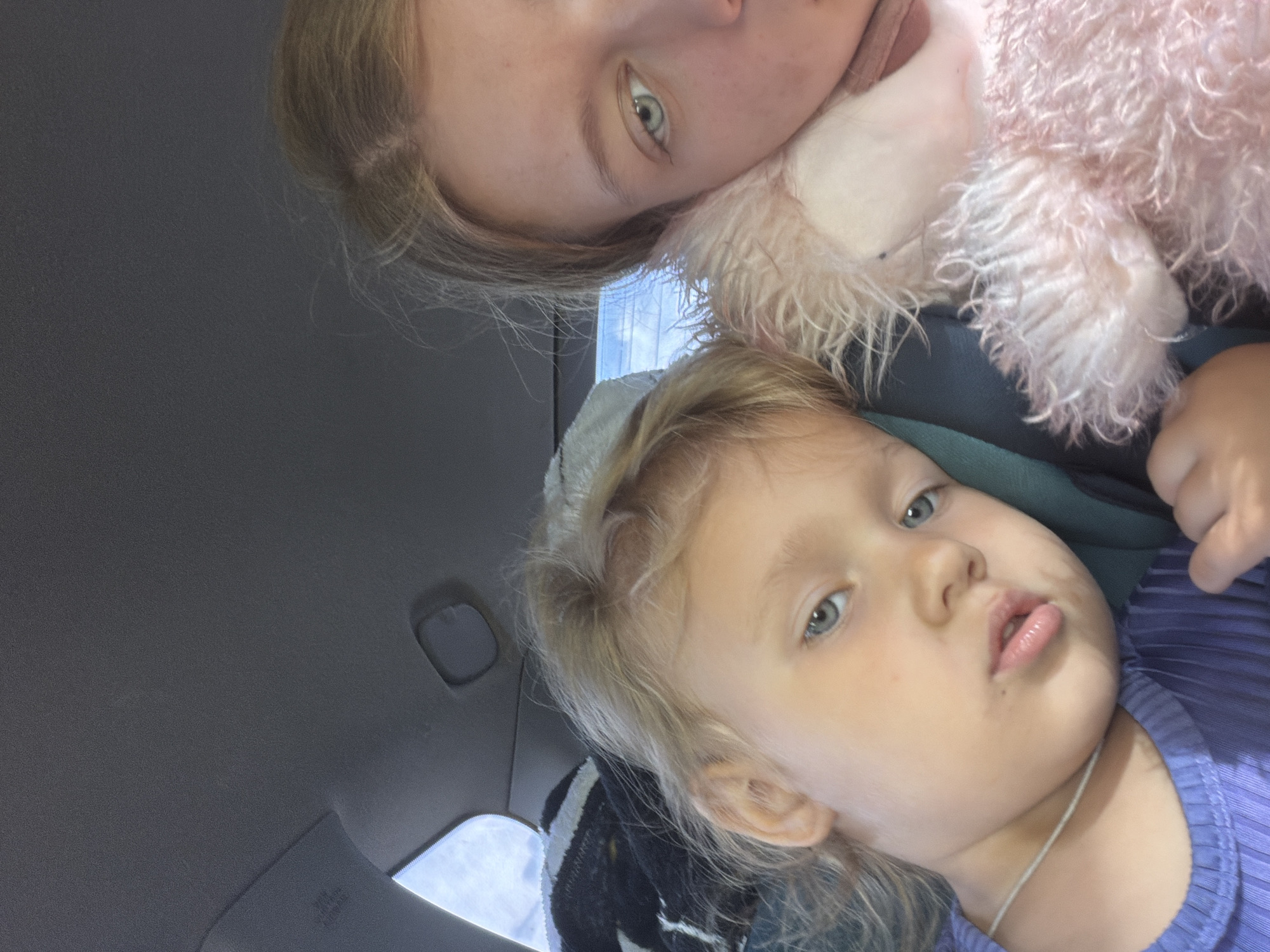 This image shows a close-up selfie of two young females inside a vehicle. On the left, a young child with blonde hair and blue eyes is visible, wearing a purple ribbed top and a thin silver necklace. Her head is tilted, and her gaze is directed slightly off-camera. To the right, a slightly older female with blonde hair is partially visible, resting her head on a fluffy pink stuffed toy. The interior of the car is dark, with hints of a gray headliner and a visible airbag label. Sunlight streams in from a window, illuminating the subjects. The scene appears to be within a moving vehicle, given the nature of the selfie and the interior context. The location in Новое Маврино, Russia, is not directly evident from the visual information within the image itself, but it is provided as contextual information.