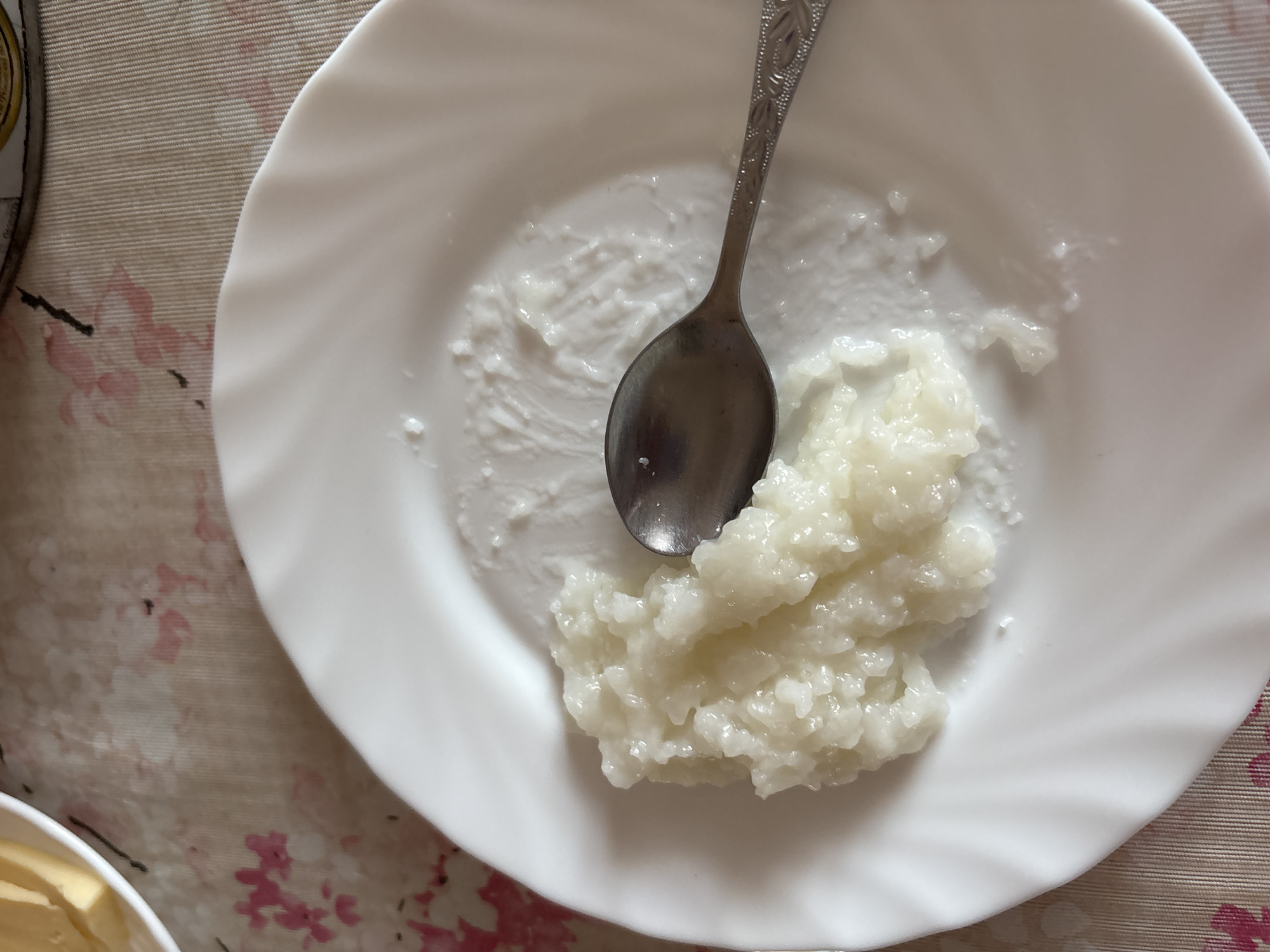 A white plate containing a pile of cooked rice and a metal spoon sits on a table in Orel, Russia. A small amount of rice is smeared on the plate's surface. The table is covered with a patterned tablecloth. Part of a dish with a yellow substance is visible in the lower-left corner of the image.
