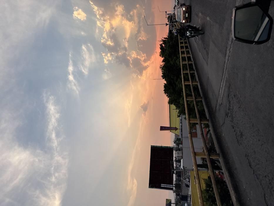 The image depicts an elevated view of an urban environment in Nezahualcóyotl, Mexico, at sunset. The sky displays a gradient from light blue with wispy clouds at the top to vibrant orange and pink hues near the horizon, with prominent crepuscular rays and illuminated cumulus clouds.
In the foreground, the dark gray asphalt surface of an elevated road or overpass is visible, with a vehicle's side mirror partially obscuring the lower right corner, indicating the photograph was taken from a vehicle. A yellow-painted concrete railing runs along the right side of the elevated structure.
On the road, a dark-colored motorcycle, ridden by a person wearing a helmet, is traveling away from the viewer. Behind the motorcycle, a line of cars is visible, including a light brown/orange sedan, also moving in the same direction.
Below and to the left of the elevated road, various buildings with flat roofs and some yellow facades are visible, partially obscured by green trees and foliage. Prominent features include two large, unlit rectangular billboards on rooftops and a tall, slender purple sign shaped like a downward-pointing arrow, bearing white text that appears to be "FRESKO" or "FRESKI". Distant urban structures and hazy hills are faintly discernible on the horizon beneath the sunset glow. Streetlights are visible but unlit.