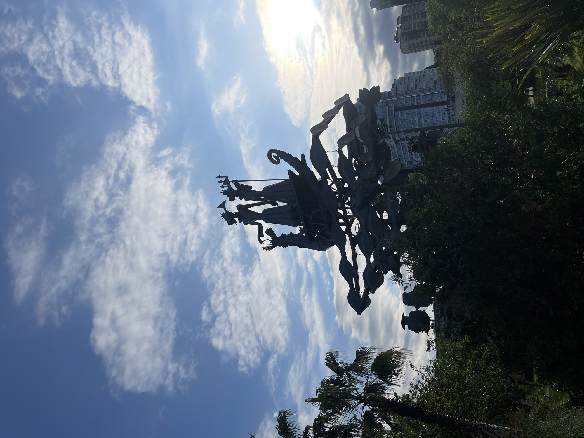 The image depicts a sculpture of the mythical Greek god Poseidon in the city of Sochi, Russia, standing in front of buildings and lush greenery against a backdrop of a blue sky with white clouds. The sculpture is silhouetted against the bright sunlight, highlighting its intricate details. In the background, modern buildings can be seen, suggesting the sculpture's location in a contemporary urban setting. The scene includes palm trees and other vegetation, indicating a park or landscaped area.