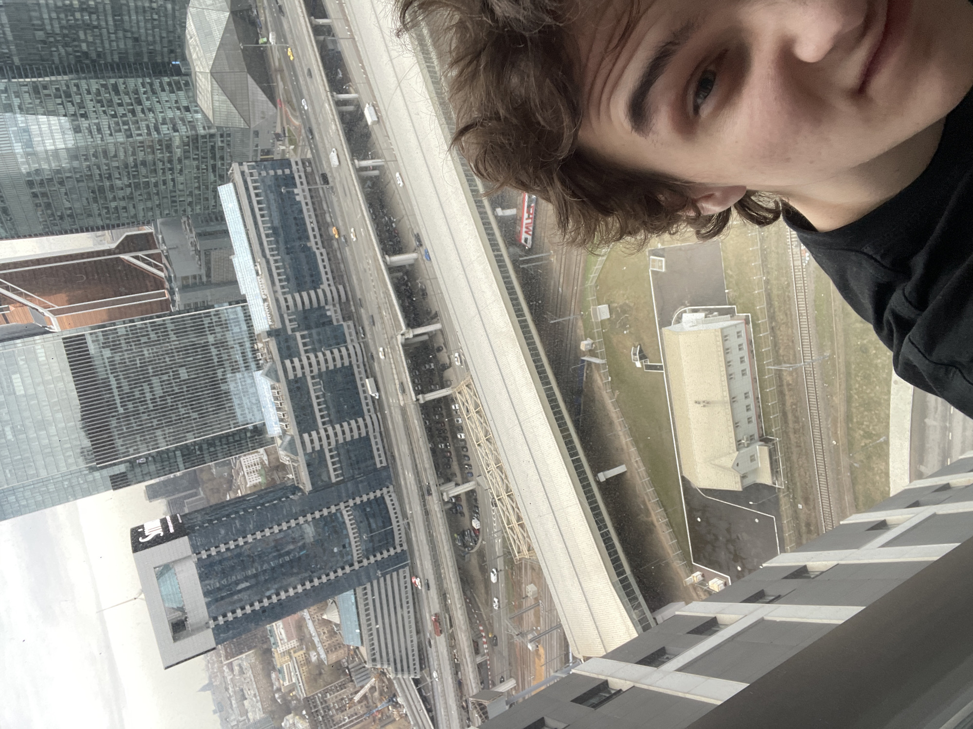 A selfie of a young man with brown, curly hair and a black shirt. The background shows a high-angle view of a dense urban landscape, likely taken from a high-rise building. Multiple modern skyscrapers are visible, along with roadways and railway tracks below. A red train is visible on the railway. In the lower right corner, part of the building the photo is taken from can be seen. The scene indicates an urban area, possibly Khimki, Russia, based on the architecture and layout of the city.