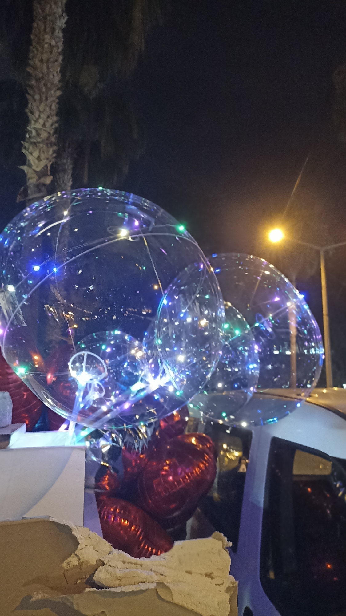 The image captures a vibrant nighttime scene featuring three large, transparent spherical balloons filled with multicolored LED lights, which glow predominantly in blue, green, purple, and white. These illuminated balloons are the central focus, creating a festive atmosphere. Below them, several red, heart-shaped foil balloons are partially visible.
The setting is outdoors at night, with a dark sky occupying most of the background. A bright yellow street light is visible in the upper right quadrant, casting a warm glow. To the far left, the textured trunk of a palm tree is visible, suggesting a warm climate. The lower right portion of the image shows part of a white vehicle, possibly a car or van, with reflections in its window, indicating the balloons might be displayed on or within it.
The overall impression is one of a celebration or event, possibly taking place on a street in Yenişehir, Turkey, given the location context and the presence of the palm tree. No people are visible, and there is no discernible text in the image.