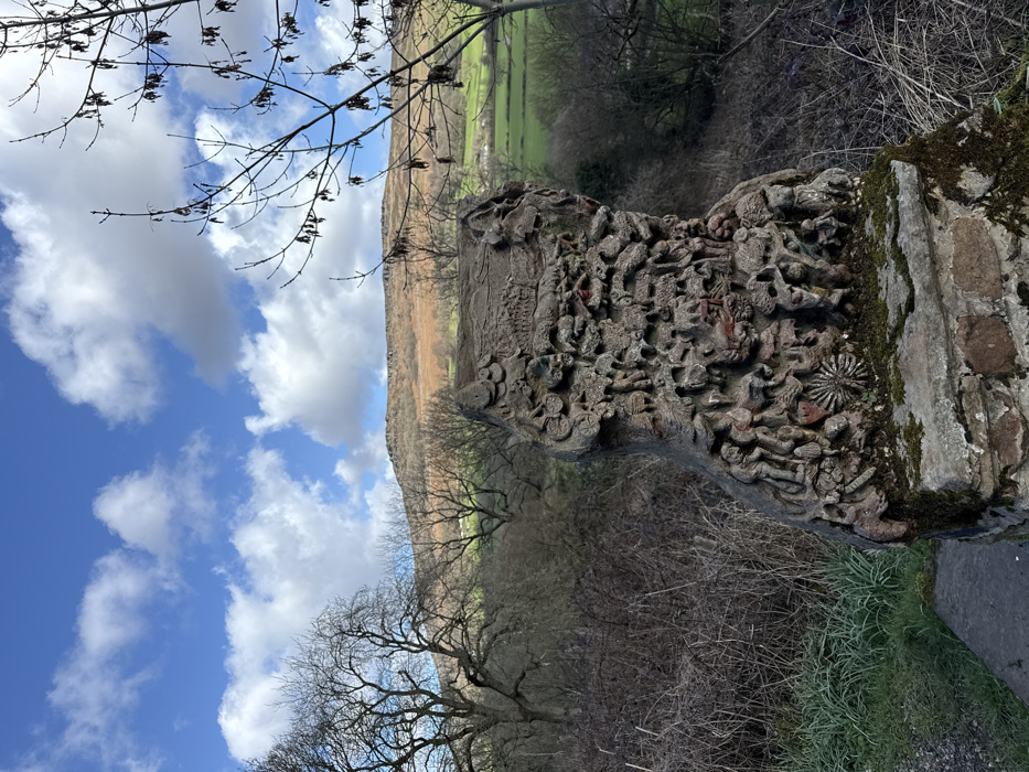 The image displays a weathered, intricately carved stone sculpture, possibly concrete, resting on a moss-covered stone and brick plinth in a rural setting within the High Peak area of the United Kingdom. The sculpture, which appears columnar, is densely decorated with numerous small, low-relief figures and patterns, exhibiting a dominant grey-brown coloration with sparse, faded red accents. To its immediate left is a narrow, light-grey path bordered by short green grass. The midground surrounding the sculpture is characterized by dense, dry, tangled brown shrubbery and several large, bare deciduous trees with dark, gnarled branches. In the background, a rolling landscape unfolds under a bright blue sky dotted with white cumulus clouds. A prominent hill or moorland covered in light brown, dry vegetation occupies the upper background, while sections of vibrant green fields and faint distant structures are visible below it. A bare tree branch with dark, dried seed pods extends into the frame from the upper right. No persons are present.