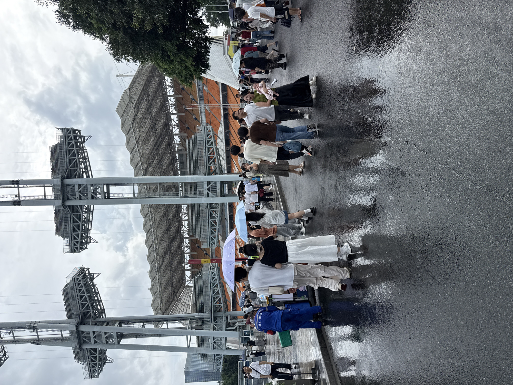 The image shows a group of people walking on a wet asphalt surface in front of a large stadium. The stadium has an orange seating area and a modern, angular roof structure, supported by massive metal pylons with platforms at the top. The sky is overcast, and puddles on the ground reflect the surrounding environment and the figures of the people. Some individuals are holding umbrellas. In the background, there are modern buildings. A worker in a blue uniform with Chinese characters on his back is visible on the left, pushing a cart. The scene appears to be located in Tianhe District, China, likely near a sports venue.
