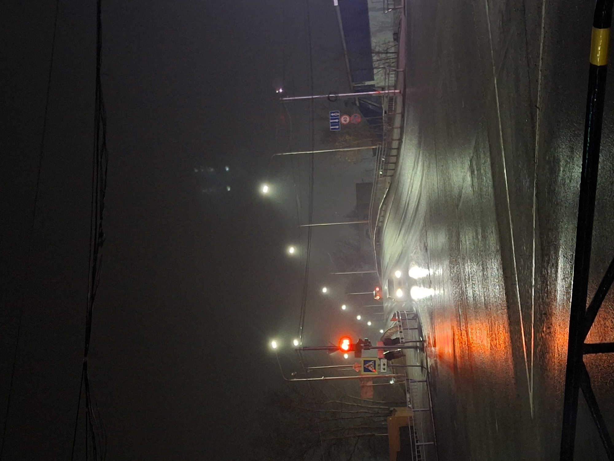 The image shows a street scene at night in what appears to be Chaikovsky, Russia. It is a foggy night, and the road is wet, reflecting the artificial light sources present. The street is illuminated by streetlights, which are spaced along the side of the road. A traffic light is visible, displaying a red signal. There are some traffic signs, and other signs that are also visible on the poles beside the road. The road surface shows signs of vehicle use. In the distance, the headlights of vehicles can be seen approaching. A set of power lines are visible on the left side of the frame. The perspective is from ground level.