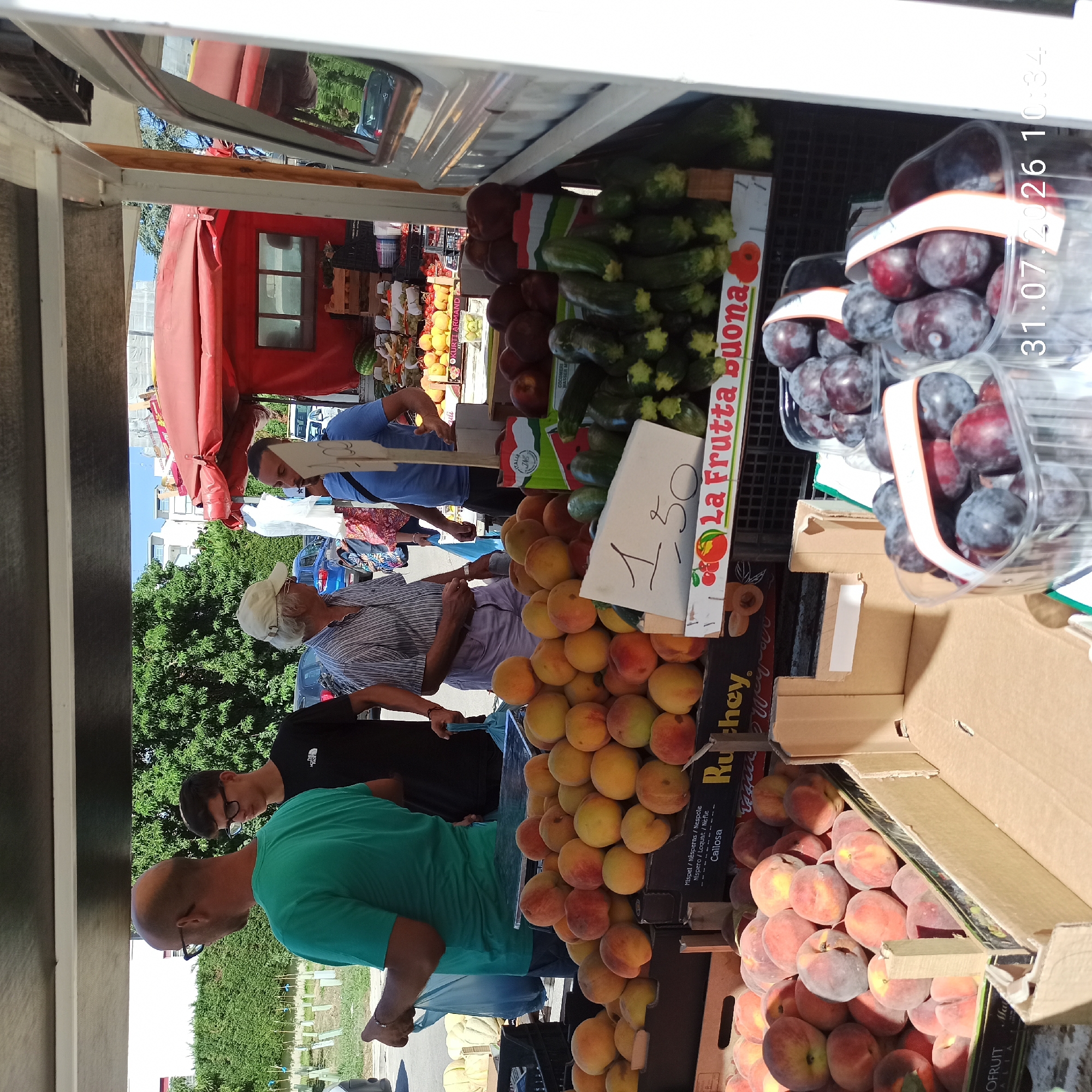 A man in a teal t-shirt and glasses is standing in front of a fruit stand and holding a blue plastic bag. To his left, a younger man wearing glasses and a black t-shirt with "The North Face" logo also stands near the stand. An older man in a striped shirt and a cap is interacting with a vendor. Several other individuals are visible in the background. The fruit stand displays various fruits including peaches, plums, and zucchini, with price tags in Italian visible on some items. The scene appears to be a market or roadside stall. The presence of a red awning and the architecture in the background suggest an outdoor market setting in Modugno, Italy, possibly near a residential area given the buildings visible.