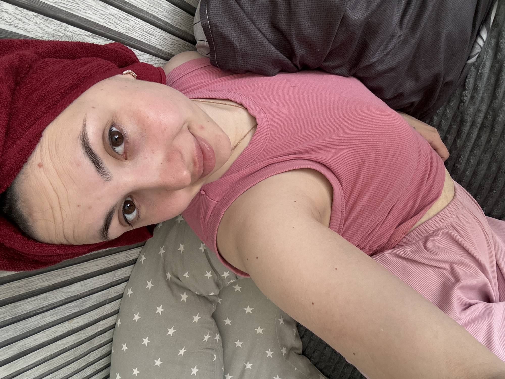 A woman lies on a grey surface, possibly a bed, in a room with striped wall or furniture detail. She is wearing a pink ribbed tank top and matching pink pants. A red towel is wrapped around her head. A grey pillow with white stars is visible beneath her. Part of a darker grey pillow is also in the frame. The location, Марковский, Russia, is extraneous to the visible content.