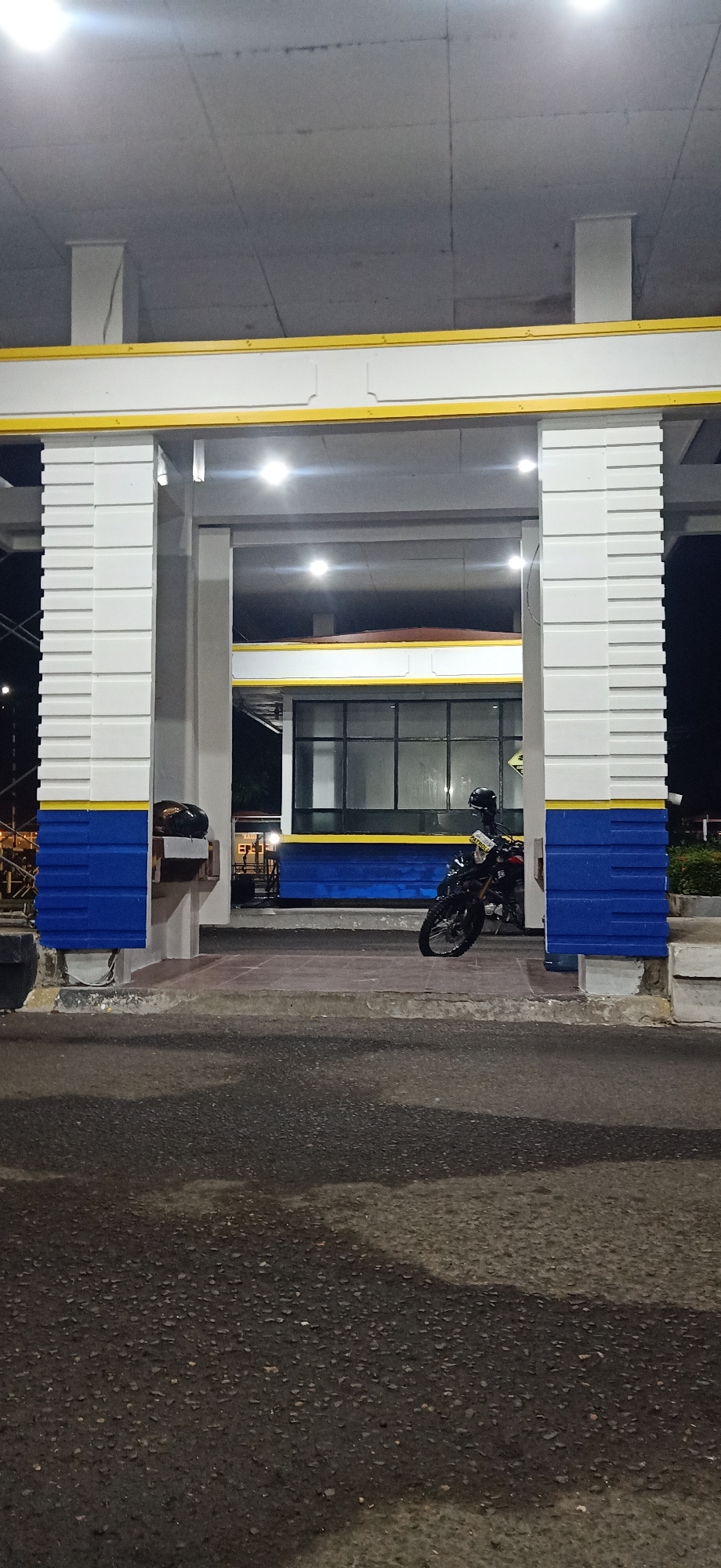 The image depicts a nighttime scene in Tambontunong, Indonesia, likely at a covered public space or entrance. The foreground is an asphalt surface with visible patches of wetness. A large, white, rectangular structure with decorative blue and yellow banding dominates the mid-ground. This structure has two prominent pillars with textured white cladding and blue banding at the base.
Within the covered area of this structure, a black motorcycle is parked. The motorcycle has a black helmet resting on its seat and appears to be positioned for easy access. Behind the motorcycle, there's a building with large windows and a darker interior. Lights are visible inside the building and also illuminate the covered area from above, casting bright circles on the ceiling. To the left of the structure, partially visible, is a black object that could be another helmet or bag. The overall impression is one of an orderly, functional space, possibly a transit point or a designated parking area, illuminated for nighttime use. The background is dark, suggesting it's nighttime in a potentially semi-urban or rural setting given the lack of extensive city lights.