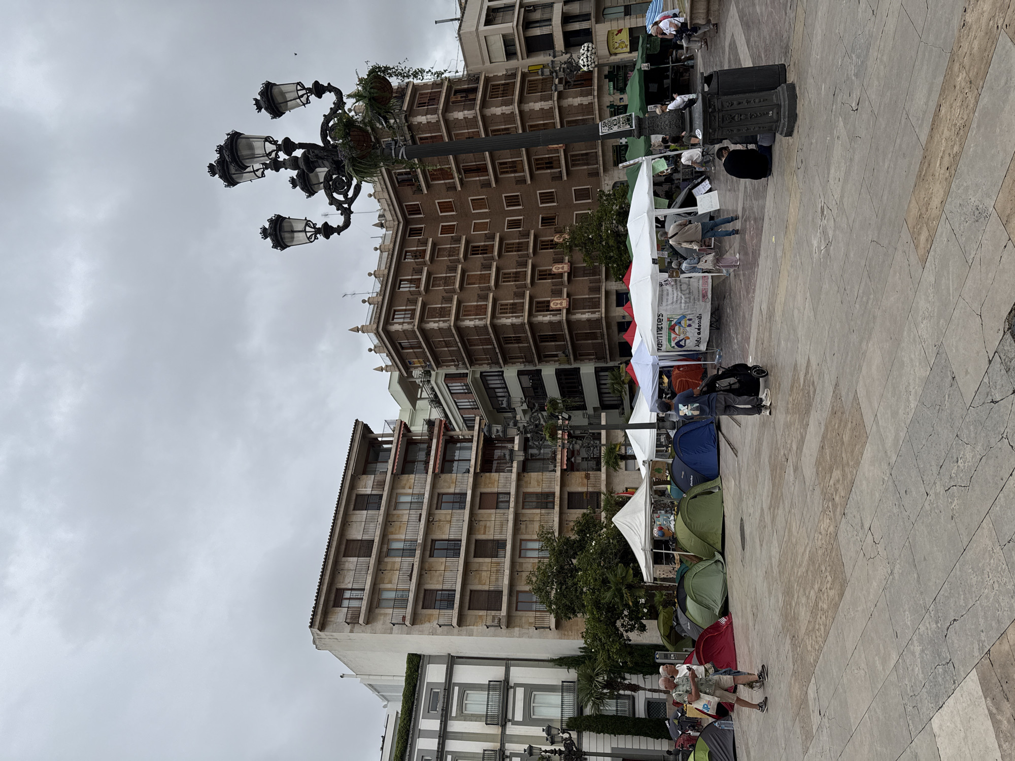 An outdoor urban scene unfolds on an overcast day, likely in a Spanish city like Valencia. The central view is dominated by several multi-story residential or commercial buildings featuring numerous windows, some with closed shutters, and projecting balconies. A large, ornate black streetlamp, adorned with some greenery, stands prominently on the left side of the frame.
The ground is a broad, light-colored stone-paved plaza or square. On the right side, a series of white market-style tents and several small camping tents in various colors (green, blue, red) are set up in rows. These tents suggest either a market, a craft fair, or potentially an encampment for a protest or public event. Several people are visible around these tents, some appearing to be browsing, others perhaps manning stalls or participating in the event. In the foreground, people walk across the paved area. A large, dark object resembling a speaker or a decorative bin is also present near one of the white stalls.
Visible text on a white banner reads "sortilegios" and "Artesania", with other partially obscured words like "Nen adesa", indicating the presence of craft goods or local products. Another sign has "P...TICKS" visible. The architecture of the buildings, with their balconies and shuttered windows, reinforces the European, specifically Mediterranean, urban setting.