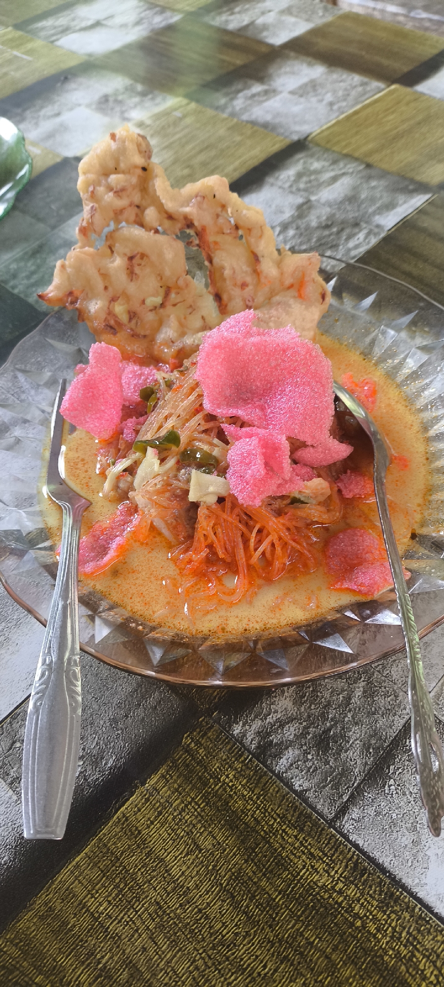 The image shows a close-up view of a dish of food, likely an Indonesian dish called "Lontong Sayur," served in a clear, faceted glass bowl. The dish consists of rice cakes (lontong), shredded vegetables, possibly chayote or papaya, in a yellowish, savory broth. It is garnished with pink crackers (krupuk) and what appear to be fried shallots or other crispy toppings. A fork is placed on the left side of the bowl, and a spoon on the right. The food is presented on a table with a patterned surface that resembles a mosaic of gray, white, and beige tiles, with a section of textured dark green fabric in the foreground. The background is also the patterned table surface. The lighting suggests it is daytime. There are no people visible in the image. The location is identified as Aluebutuha, Indonesia.