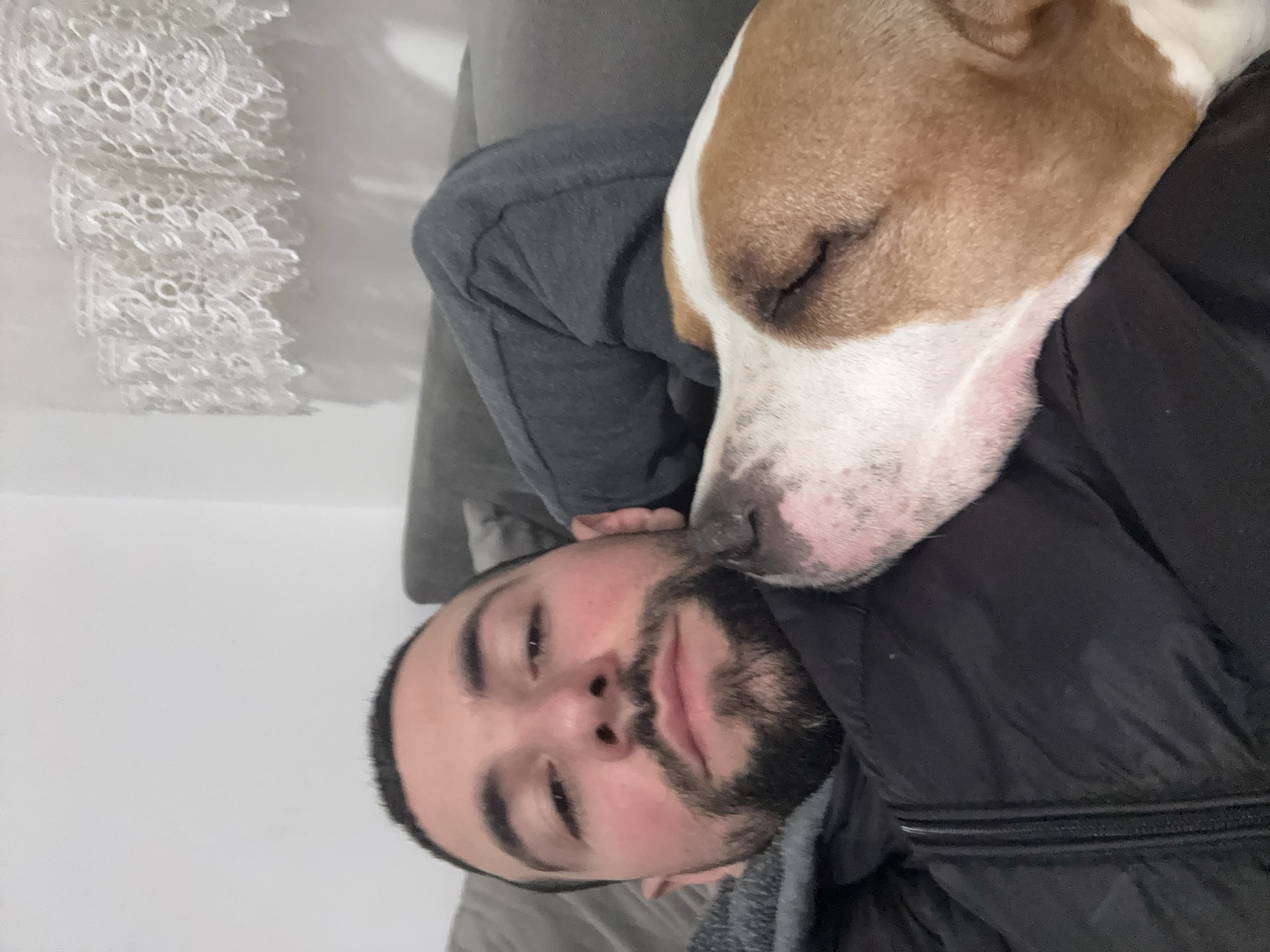 A man is lying on a gray couch with a brown and white dog resting its head on his chest. The man has dark hair, a beard, and a light complexion. He is wearing a gray hooded sweatshirt and a black jacket. The dog's eyes are closed. A white lace-like object is visible in the background. It is not possible to determine the location as Sofia, Bulgaria from the image provided.