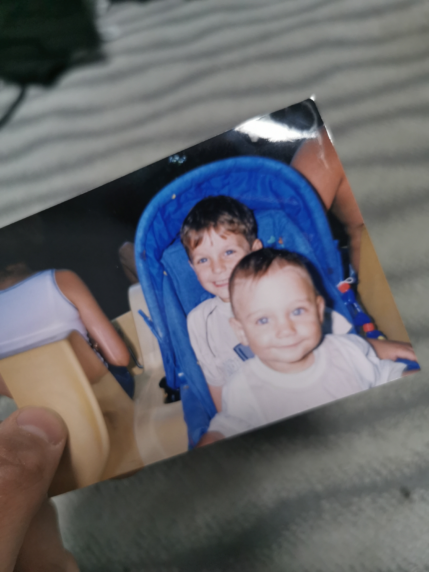 A color photograph held by a person features two young boys in a blue stroller. The older boy has short brown hair, while the younger boy has lighter hair and blue eyes. Both are wearing white shirts. The photo appears to be taken indoors or in a shaded area. There is no indication of the photo having been taken in Varna, Bulgaria.