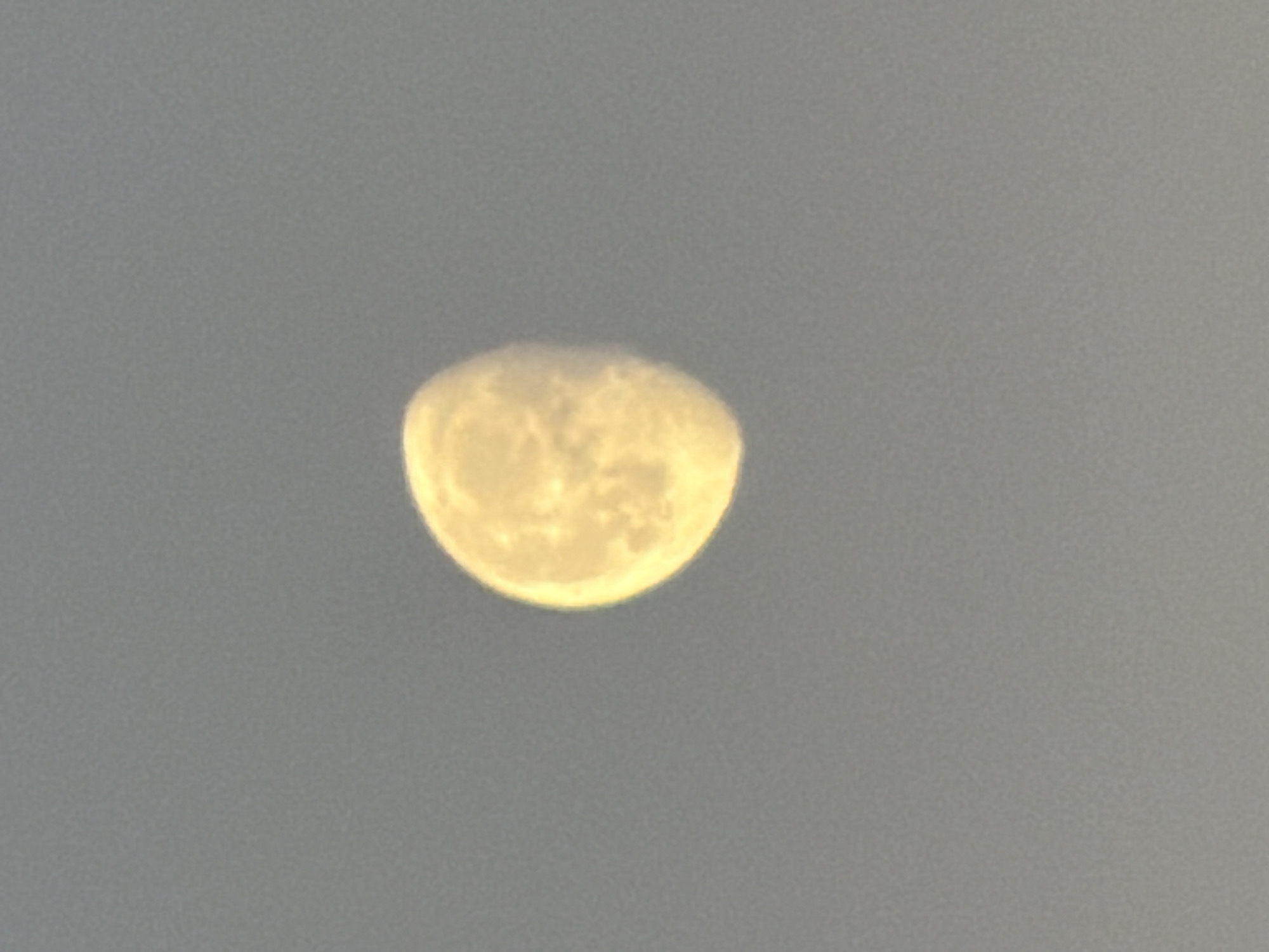 The image shows a bright, gibbous moon in a hazy, light gray sky. The moon appears slightly distorted from a perfect circle. The location in Leudelange, Luxembourg, is not discernible from the image. There are no people, subjects, objects, actions, or interactions visible other than the moon itself in the sky. The environment consists of a cloudy or hazy atmosphere.