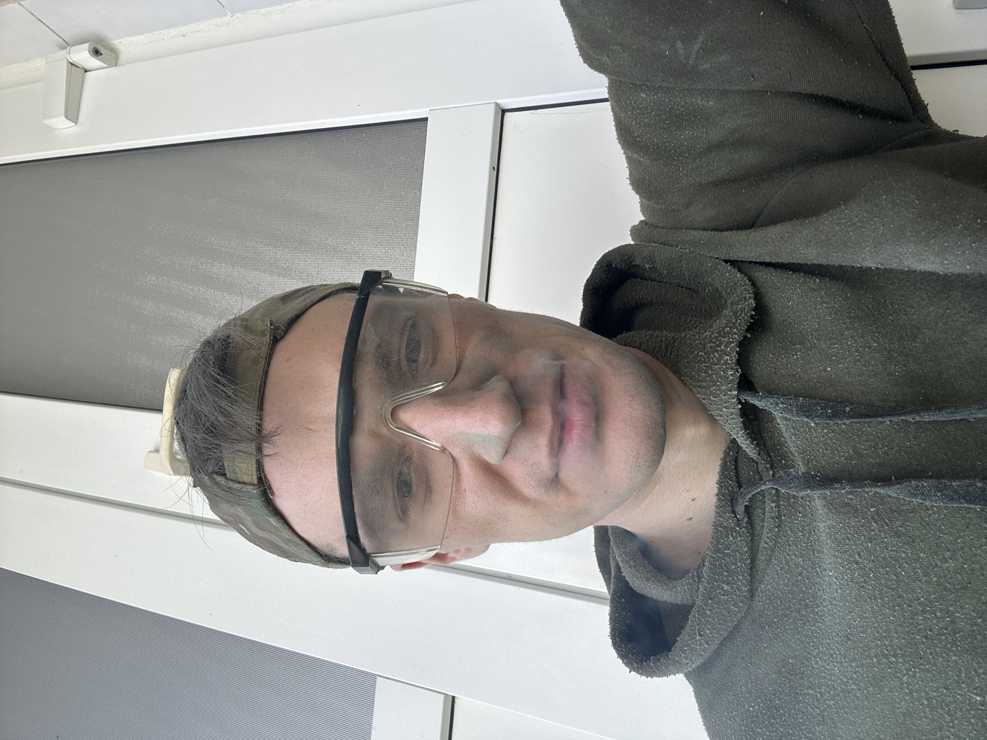 A man wearing a camouflage cap, safety glasses, and a dark green hooded sweatshirt stands in front of a white door with a screen. He is looking directly at the camera and appears to be smoking. The background consists of the white door and part of a wall. The image was taken outside, with natural light. The location is Makiv, Ukraine.