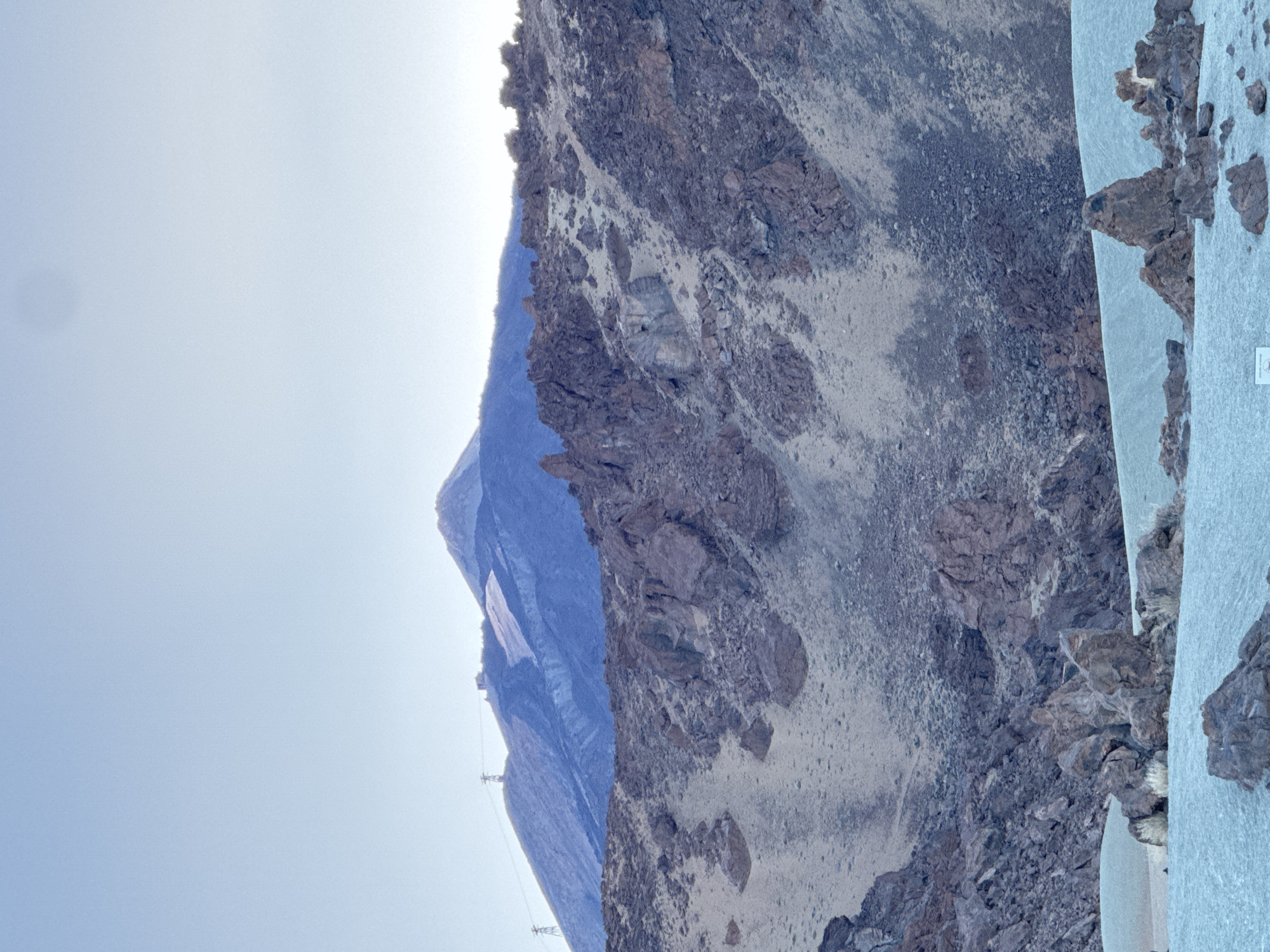 The image shows a mountain landscape under a clear sky. In the foreground, there are rocky, barren slopes with sparse vegetation. In the mid-ground, the terrain is also rocky and uneven, with patches of lighter-colored soil or sand. In the background, a large, imposing mountain dominates the scene, with a snow-capped or frost-covered peak. Two tall, slender structures, resembling power line pylons, are visible on the slopes of the background mountain, along with some buildings at the summit. The overall atmosphere is one of rugged, natural beauty.