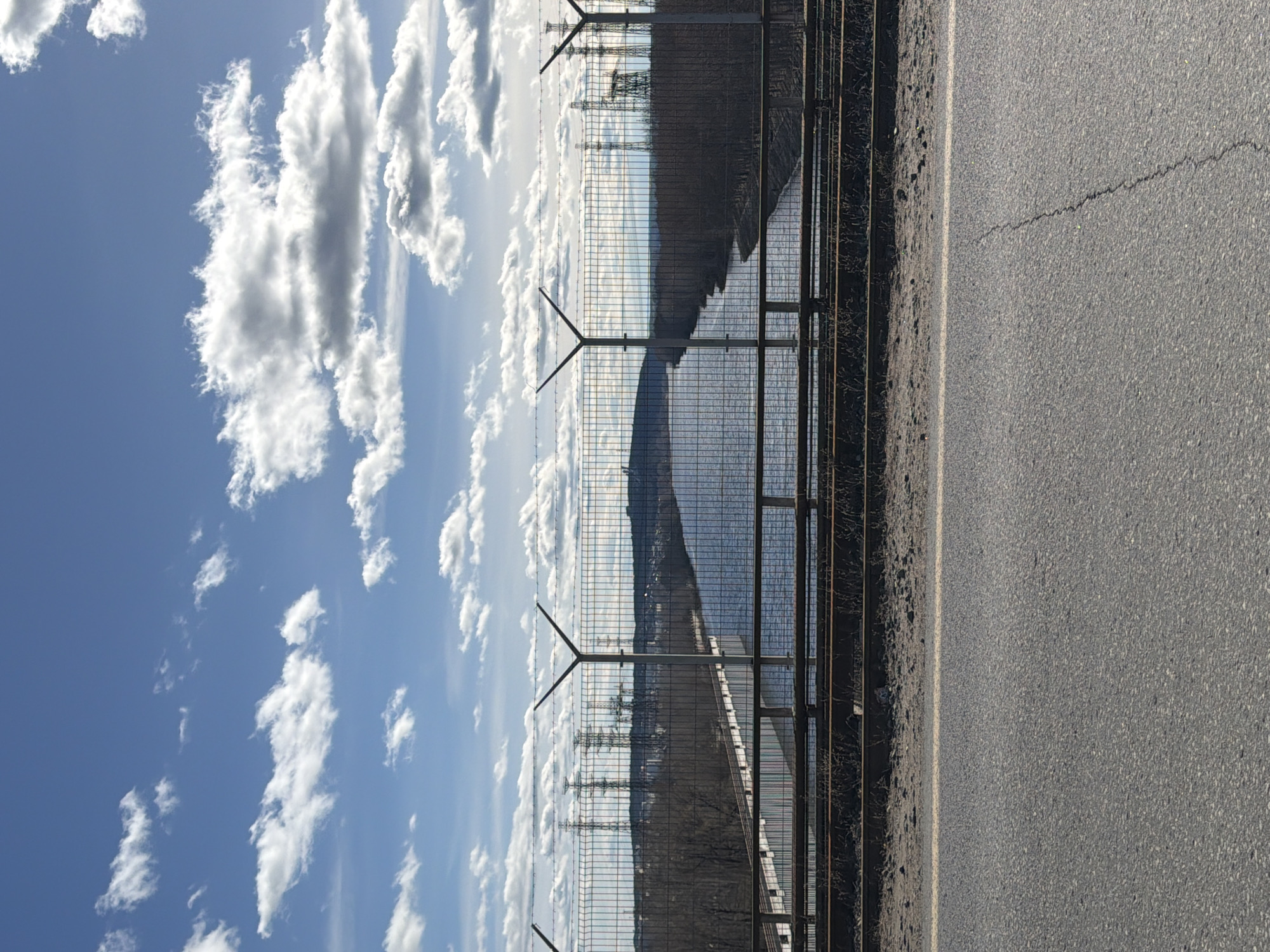 The image shows a portion of a road in Chaikovsky, Russia. A metal fence runs along the road's edge, and behind the fence, there is a body of water, possibly a river or lake. Beyond the water, power transmission towers are visible. The sky is blue with scattered cumulus clouds. The road surface is asphalt and shows some signs of wear. There are no people visible in the image.