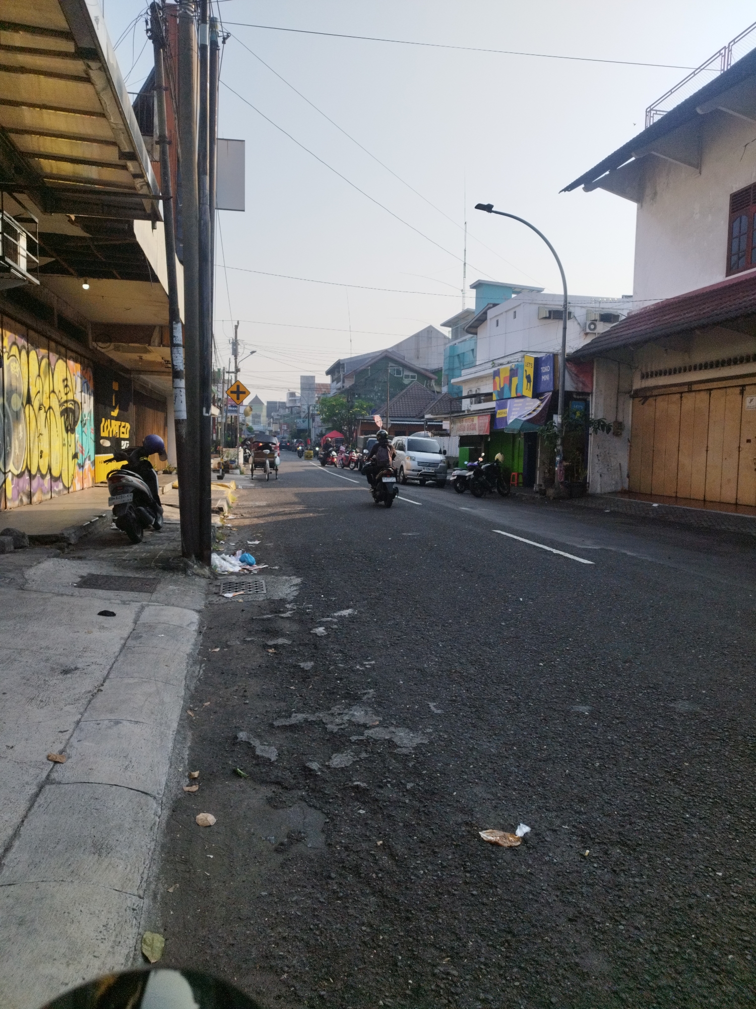 The image depicts a street scene in Yogyakarta, Indonesia, likely during daytime given the bright sky and shadows. On the left, a row of buildings features closed storefronts with metal roll-up doors adorned with yellow graffiti. A sidewalk runs along these buildings, with a parked motorcycle visible.
The street itself is paved with asphalt and shows signs of wear. Several other motorcycles are present, with some individuals riding them. In the distance, traffic signals and various shops line the street, suggesting a commercial area. The architecture of the buildings is diverse, with some appearing older and others more modern. Utility poles with numerous wires are a prominent feature of the urban landscape. The overall impression is of an active, albeit somewhat weathered, street in an Indonesian city.