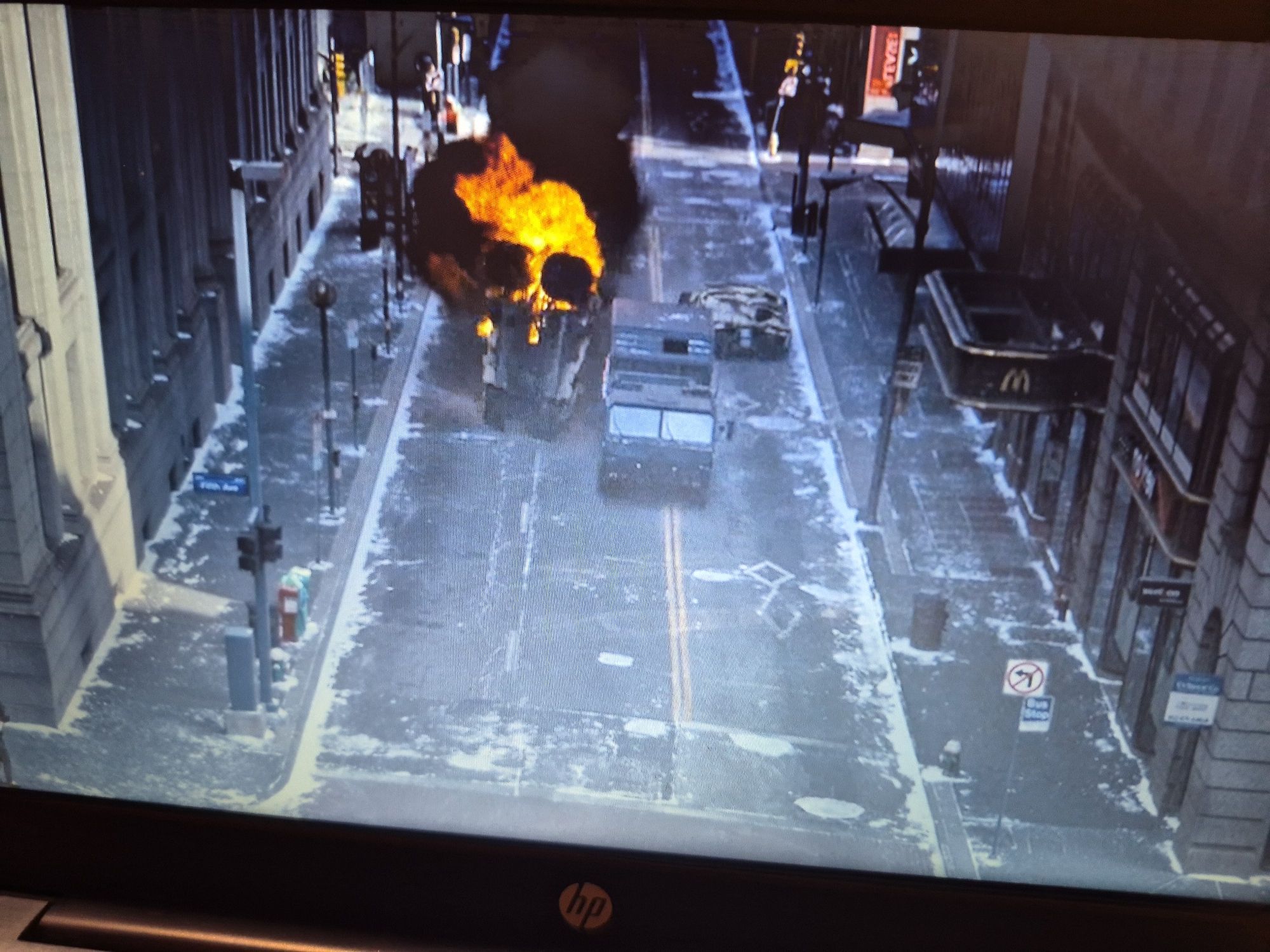 The image shows a city street scene with snow on the ground. There is a vehicle on fire in the middle of the street, with a large plume of smoke rising from it. Another vehicle, possibly a military truck, is positioned behind the burning vehicle. Buildings line both sides of the street, with some signs visible. The scene appears to be a dramatic event occurring in an urban environment. The location is identified as Sandwell, United Kingdom.