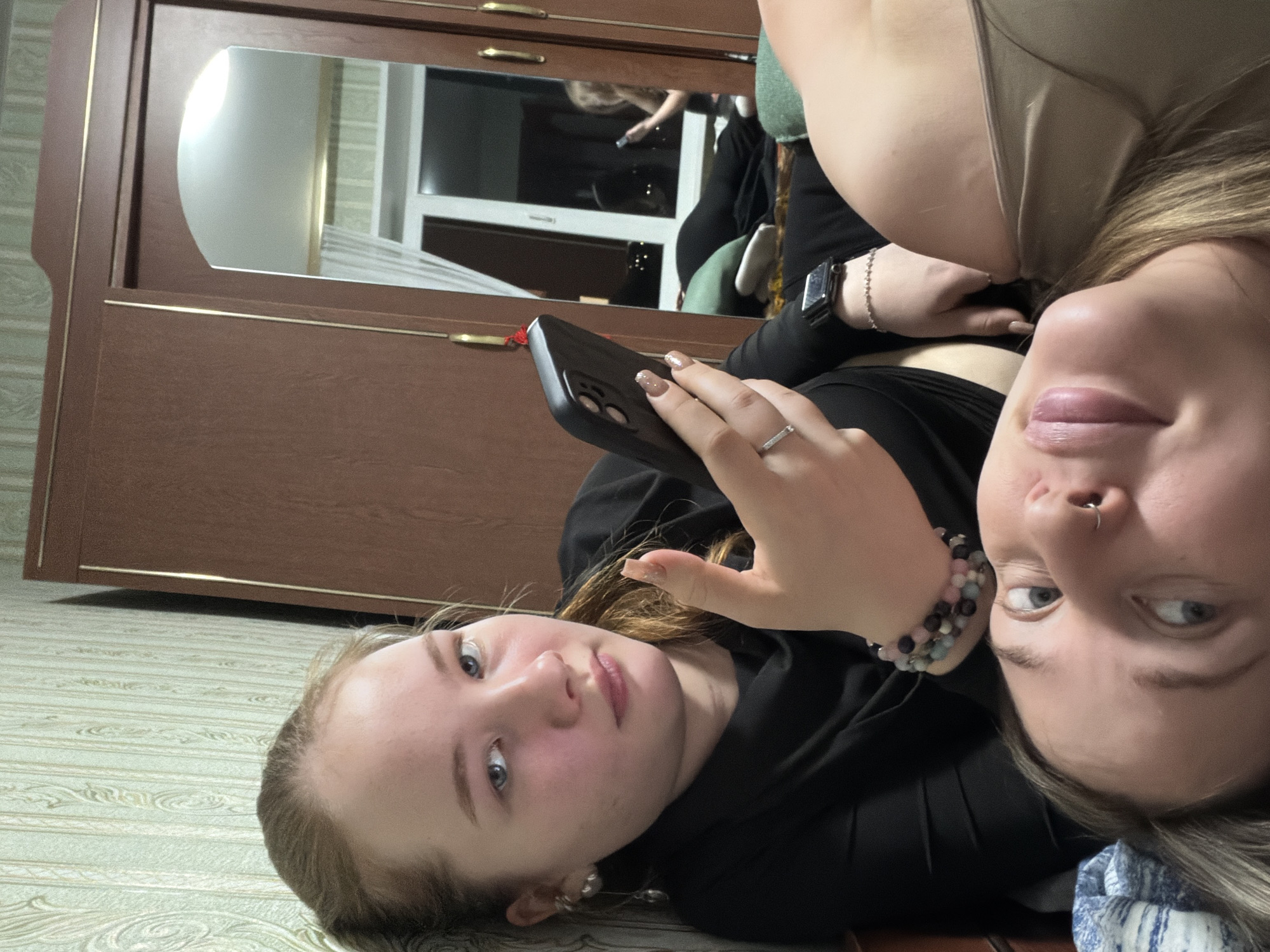 Two young women are posing for a selfie indoors. The woman on the left holds a dark-colored smartphone, showing manicured fingernails. She is wearing a black hoodie. The woman on the right has a nose ring, a beaded bracelet, and a wristwatch. A light-colored garment is visible on her shoulder. A large wooden wardrobe with a mirror is in the background. The reflection in the mirror shows more of the room, with a window and other items. The floor is a light color. Based on prior knowledge, Chaikovsky, Russia, is the nearest city.