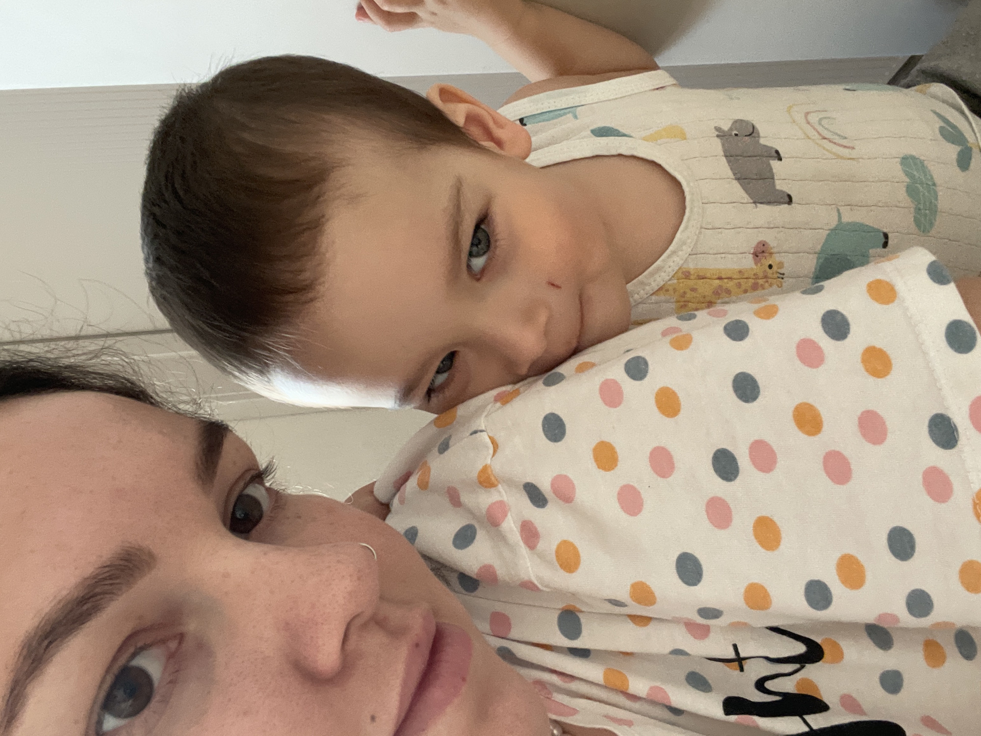 A selfie shows an adult female from the chest up and a toddler, likely her child, who is lying on her shoulder. The woman has fair skin, brown hair, and brown eyes. She is wearing a white shirt with blue, orange, and pink polka dots. The toddler has fair skin, brown hair, and light-colored eyes. He is wearing a white tank top with cartoon animal patterns. The background appears to be a plain, white wall. There is no evidence to suggest that the image was taken in Izhevsk, Russia, or any other specific location.