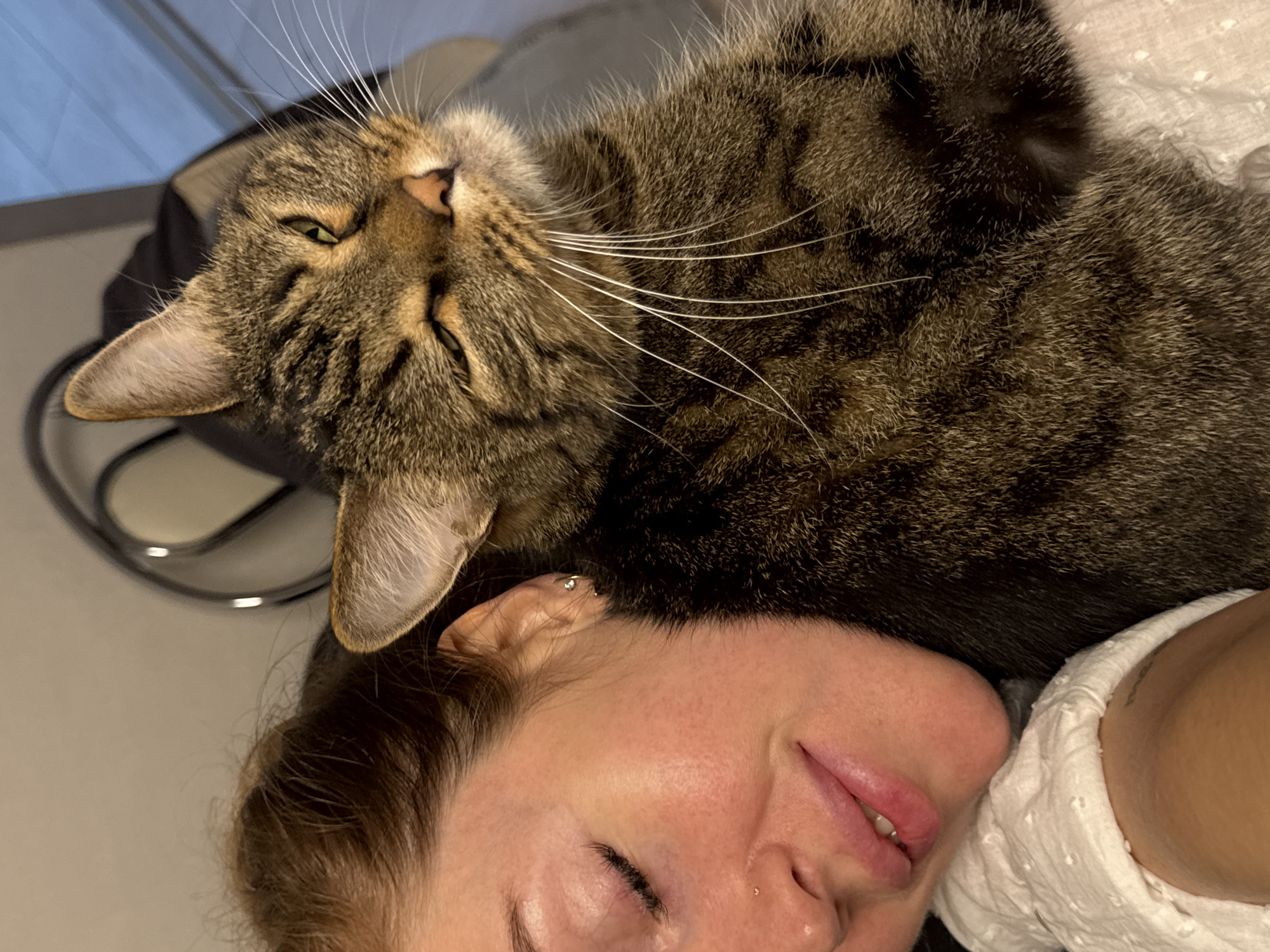 A woman lies on her back with her eyes closed. A tabby cat is lying on top of her, with its head resting on her forehead. The cat's whiskers are prominent. The scene appears to be indoors, possibly in a home. There is no visible evidence to confirm the location of Chaikovsky, Russia.