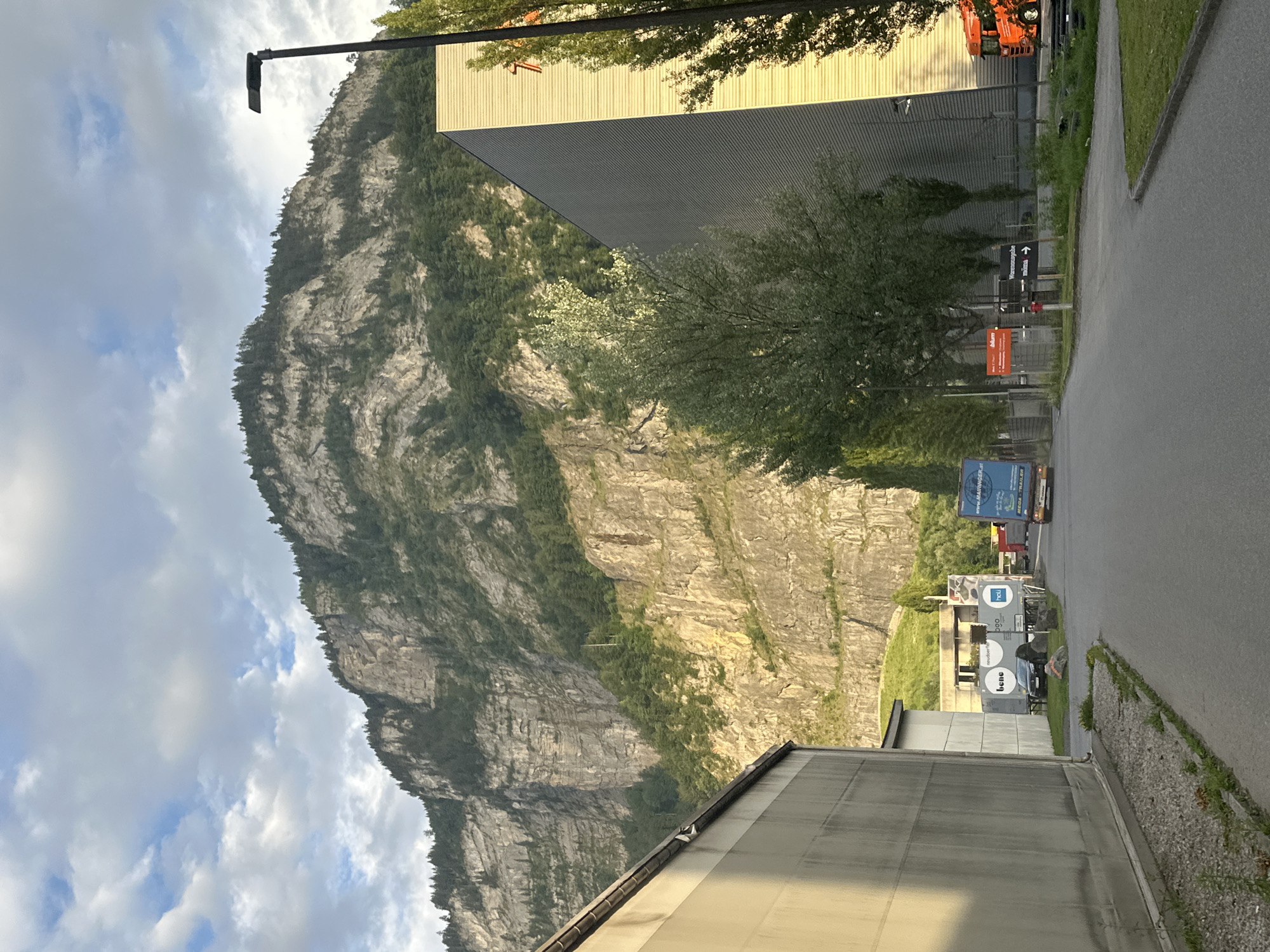 The image shows an industrial area at the base of a large, steep, rocky mountain. In the foreground, a paved road leads into the scene, bordered by a gravel path and grass. On the left, a light-colored, elongated building with a light grey facade dominates the left side of the image. On the right, a tall, modern building with a ribbed, metallic facade, predominantly light yellow in color, rises up and is partially illuminated by sunlight. A street lamp is visible on the right edge of the image.
In the middle ground, several trucks and trailers are parked or in motion. One blue truck with "MEGA TRAILER" printed on its side is visible on the right side of the road. There are also white trailers with logos like "bene" and "hau". A dark car is parked behind one of the trailers.
The background features the impressive mountain face, characterized by its rugged, grey rock formations and dense green vegetation, primarily trees, covering its slopes. The sky above is partly cloudy with patches of blue. The overall scene suggests an operational industrial zone located in a mountainous region, possibly on the outskirts of Dornbirn, Austria, given the context of the provided location.