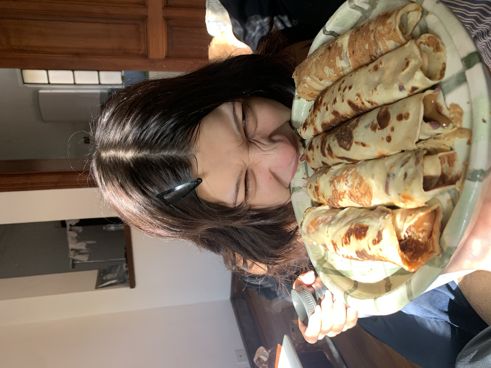A young person with long, dark hair and a black barrette is positioned close to a full plate of rolled crepes. The person is attempting to eat a crepe directly from the plate, their face contorted in a grimace, eyes closed. They are holding a small metallic object in their other hand. The plate has a green and white patterned rim and is filled with browned, rolled crepes, some revealing filling inside. The background suggests an interior setting with a dark wooden door, a window with visible light, and a cabinet. The image is taken indoors with natural lighting and appears to be rotated counter-clockwise. The location, Buenos Aires, Argentina, cannot be determined from the provided information.