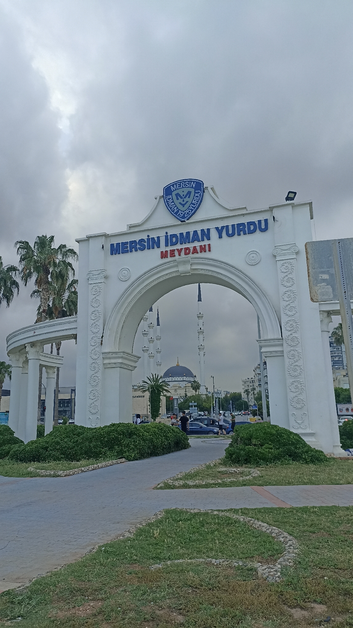 The image captures the Mersin İdman Yurdu Meydanı entrance in Yenişehir, Turkey, under a cloudy sky. The white arched gate, adorned with the text "MERSİN İDMAN YURDU MEYDANI" in blue, frames a view of the Muğdat Mosque with its distinctive blue dome and minarets. Palm trees and other greenery flank the entrance. Cars and pedestrians are visible in the background. The foreground features a paved pathway surrounded by patches of grass and shrubbery.