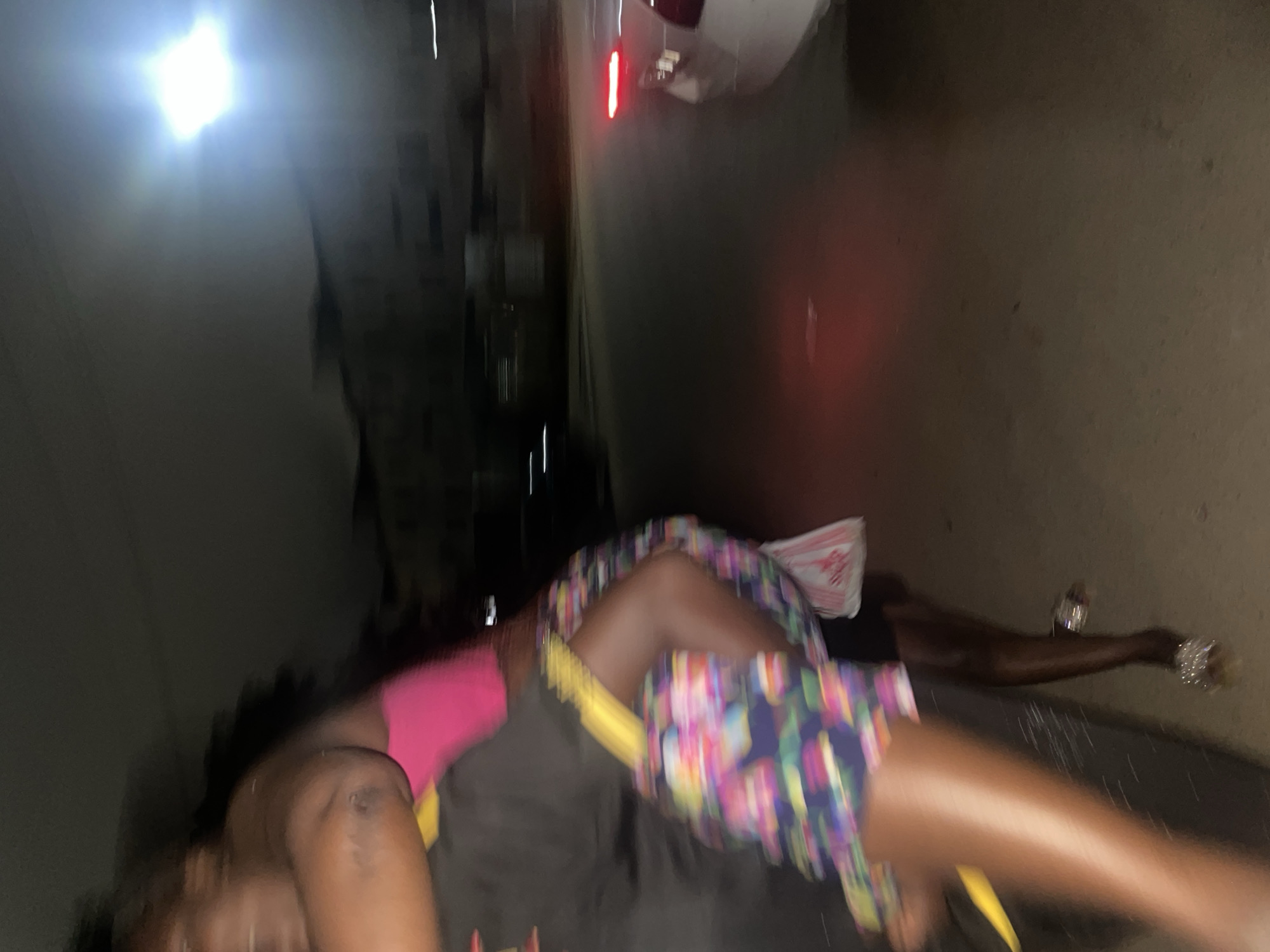 The image depicts a night scene in Guzape, Nigeria. In the foreground, a person is being carried, with their legs prominently visible. They are wearing a colorful, patterned skirt and a pink top. The person carrying them is largely obscured, wearing a black garment. Their interaction suggests a moment of support or transportation.
The environment is a street at night, characterized by blurred motion and artificial lighting. A white car with illuminated red taillights is visible on the right side of the frame, suggesting movement or stationary presence on the road. Streetlights and building lights are faintly visible in the background, contributing to the overall nighttime ambiance. The ground appears to be paved, consistent with a street surface. The overall impression is one of candid movement captured during a nighttime urban setting.