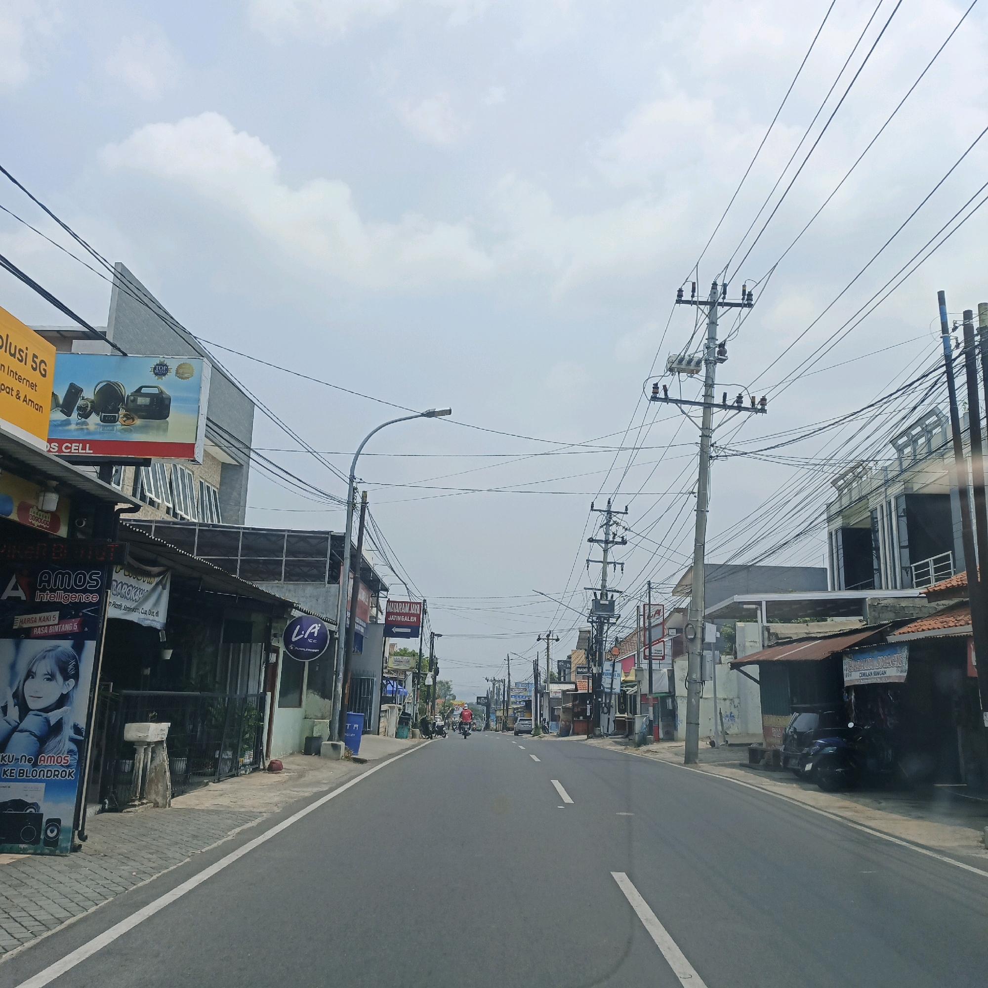 The image shows a street scene in a city, possibly in Indonesia, with buildings lining both sides of a paved road. The sky is overcast with clouds. On the left side of the road, there are several shops with signs, including one advertising "Solusi 5G" and another with an image of a woman and text that reads "KU-NE AMOS KE BLONDROK." Further down the street on the left, there is a sign for "LA ICE PURPLE HEART." On the right side of the road, there are also shops and buildings, some with orange tiled roofs. Power lines are strung across the street and supported by utility poles. A motorcycle is visible in the distance on the road, and a car is parked in front of a shop on the right. The overall impression is of a commercial street with everyday activity.
