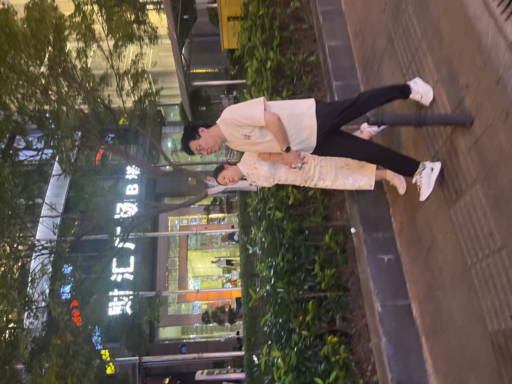 A man and a woman are walking side-by-side on a sidewalk in front of a building at night. The building has large glass doors and windows, with illuminated signage that reads "财富广场" and "B塔" in Chinese characters. The woman is wearing a light-colored, patterned dress, and the man is wearing a beige t-shirt and black pants. They are holding hands, and the man has a watch on his left wrist. The sidewalk is paved and has tactile paving in some areas. A dark-colored metal pole is positioned between them. Behind them, a manicured hedge separates the sidewalk from the building. Trees with green foliage are visible in front of the building and to the right of the scene. The lighting suggests an urban environment in Yuzhong District, China.