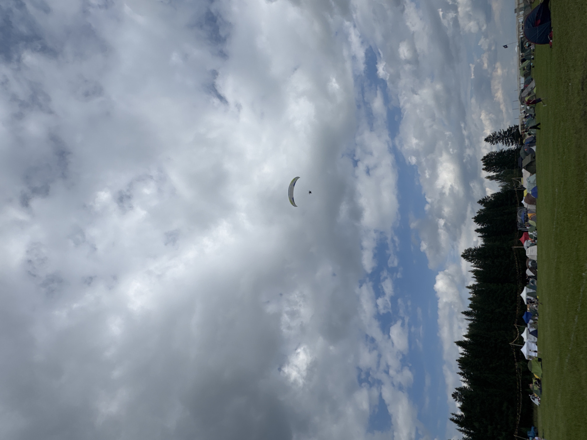 The image shows a wide view of a grassy field with a dense forest in the background. Several tents are set up across the field, suggesting a campsite or an outdoor event. In the sky, a paraglider is visible against a backdrop of a cloudy sky. The overall impression is that of an outdoor recreational or festival setting in a rural or natural environment.