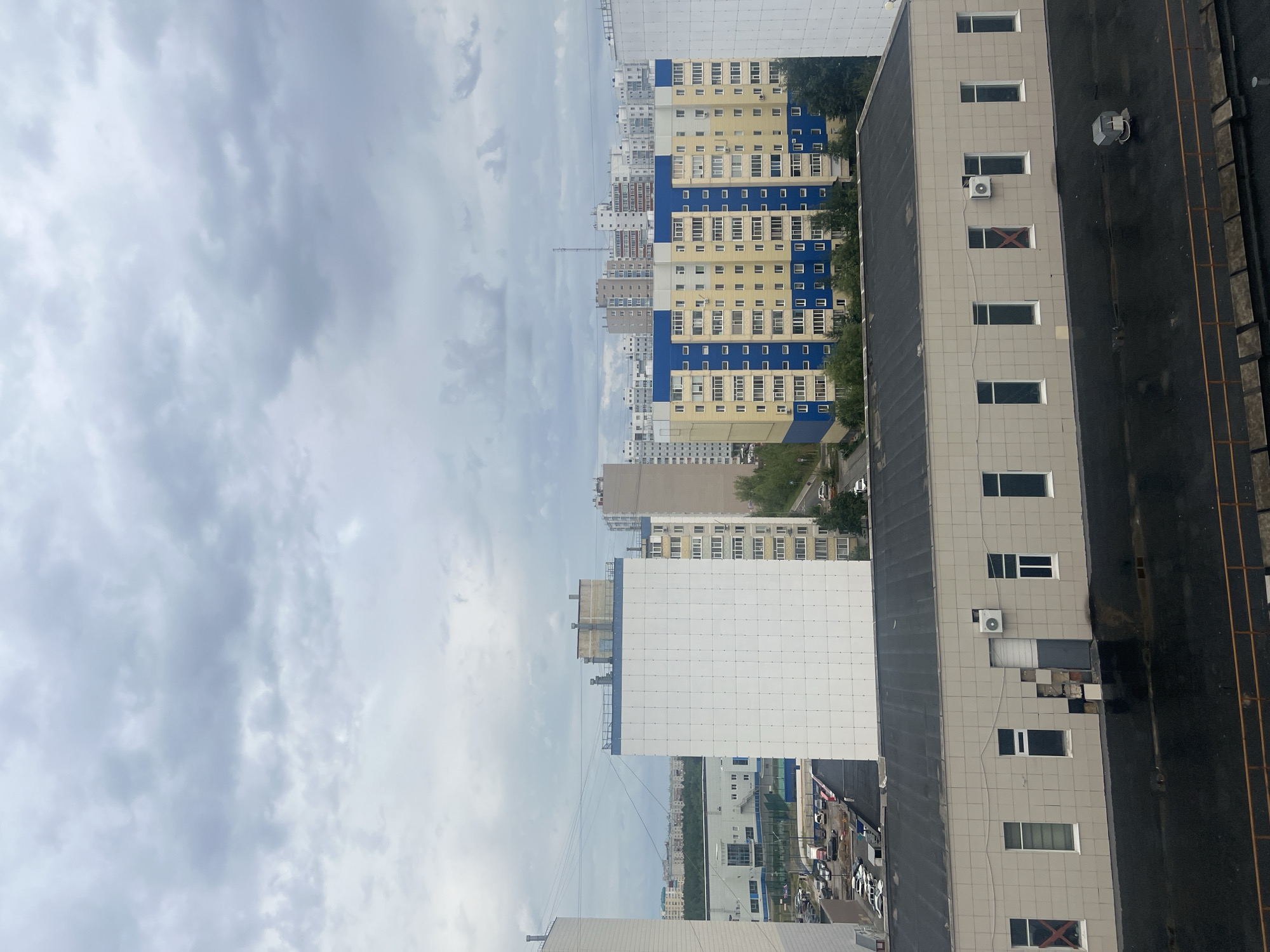 The image captures a view of an urban environment under a cloudy sky, likely in Surgut, Russia. In the foreground, a flat-roofed, light-colored building with several windows and an air conditioning unit is visible. The roof appears wet, suggesting recent rain. In the mid-ground, a large, white, multi-story residential building with a grid-like pattern on its facade dominates the scene. To its right, a yellow and blue apartment building with numerous windows stands out. Further in the background, more residential buildings of various heights and colors are scattered across the landscape, interspersed with trees and greenery. A parking lot with several cars is visible to the left of the white building. Power lines crisscross the sky. The overall atmosphere is overcast and somewhat muted due to the cloudy weather. There are no people visible in the image.
