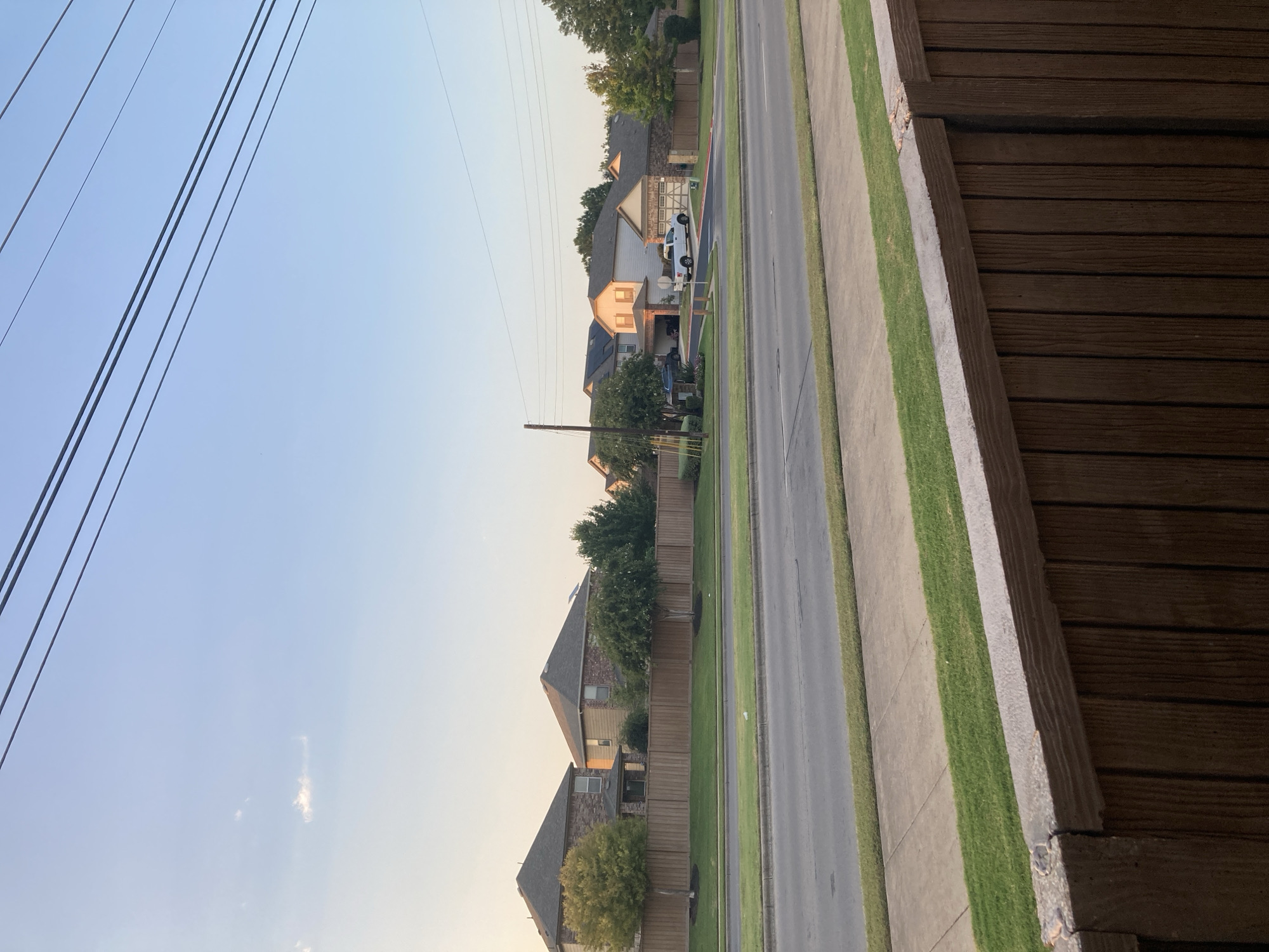 In the city of Round Rock, United States of America, a residential street is depicted with houses lining the right side of the frame. The houses are of various architectural styles, featuring brick and siding exteriors, and are separated by wooden fences. Several trees and manicured lawns are visible in front of the houses. A white pickup truck is parked in a driveway in front of one of the houses. The street itself is paved and appears to be a two-lane road with a sidewalk and a grassy median. Overhead, power lines stretch across the clear blue sky. In the foreground, a wooden fence is partially visible, suggesting the vantage point is from a yard or porch. The scene appears to be captured during the late afternoon or early evening, given the soft, warm light illuminating the houses and landscape.