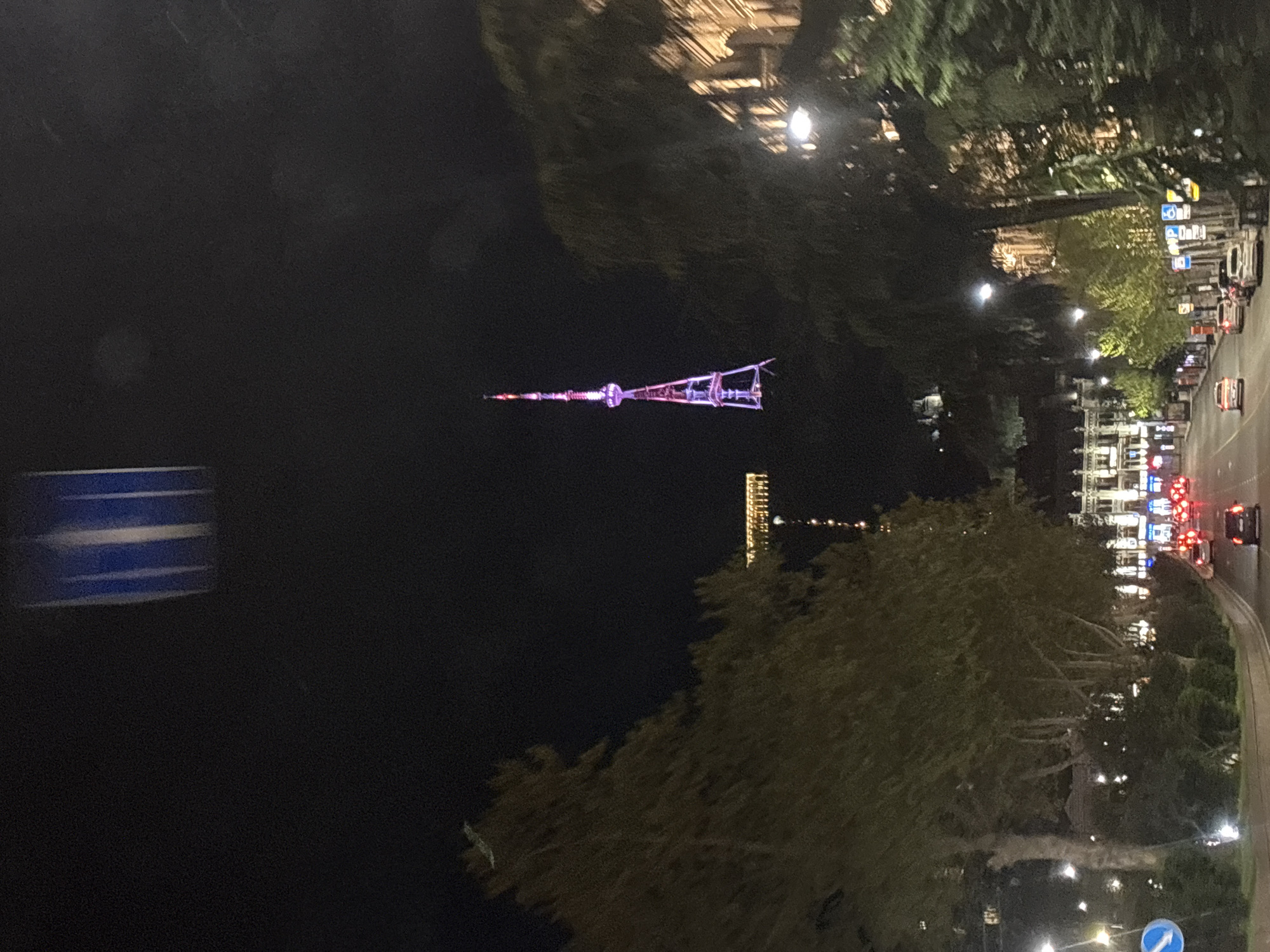At night, the image showcases the Tbilisi TV Broadcasting Tower in Tbilisi, Georgia. The tower is lit with pink and white lights and positioned in the center of the frame, appearing distant. The environment surrounding the tower is dark, punctuated by city lights, some buildings, and trees. A road with traffic can be seen on the right side of the frame. A blurred road sign is visible on the left side of the image.