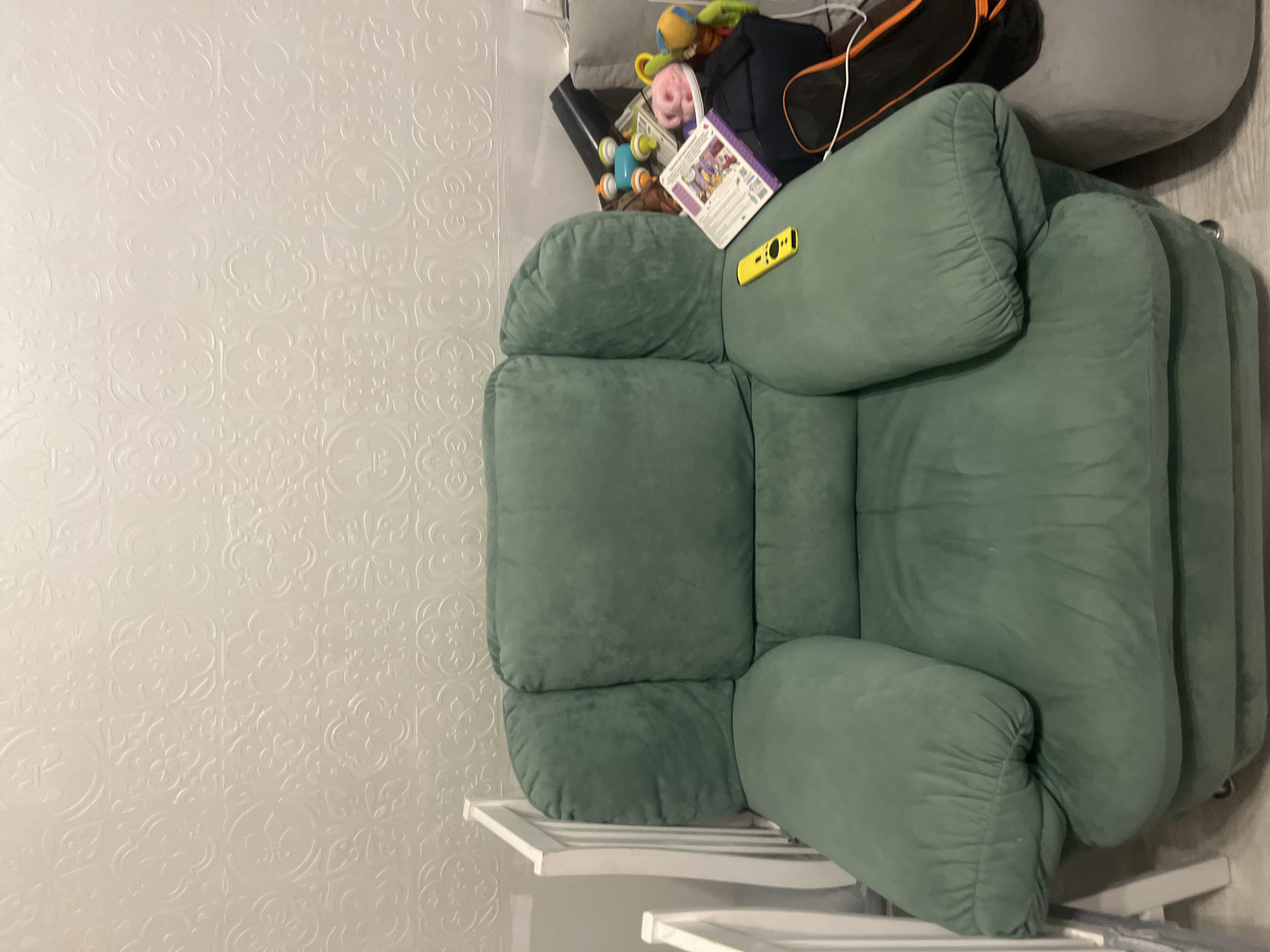 The image shows a green armchair positioned in front of a textured, light-colored wall. To the left of the armchair, a white crib is partially visible. On the right side of the armchair, several items are clustered together, including a book, a black bag with an orange trim, and various children's toys such as a pink stuffed animal, a blue and orange toy, and a yellow remote control placed on the armchair's armrest. The flooring appears to be wood. The scene is indoors, suggesting a residential setting. The location is specified as Veteran-2, Russia.