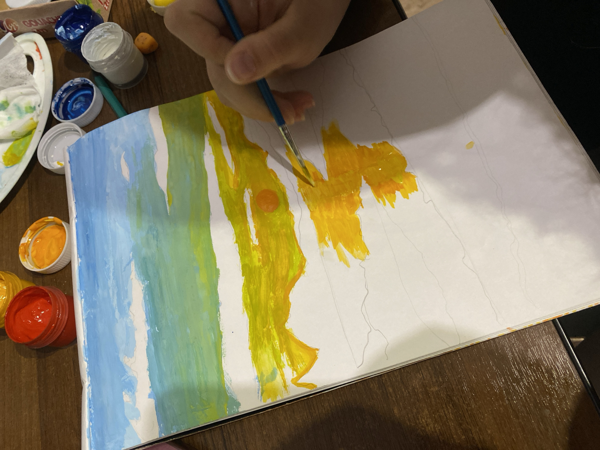 A hand holds a paintbrush applying yellow paint to a piece of paper with a drawing on it. The painting includes blue, green, and yellow colors. The painting appears to be on a table where jars of paint are resting next to it.