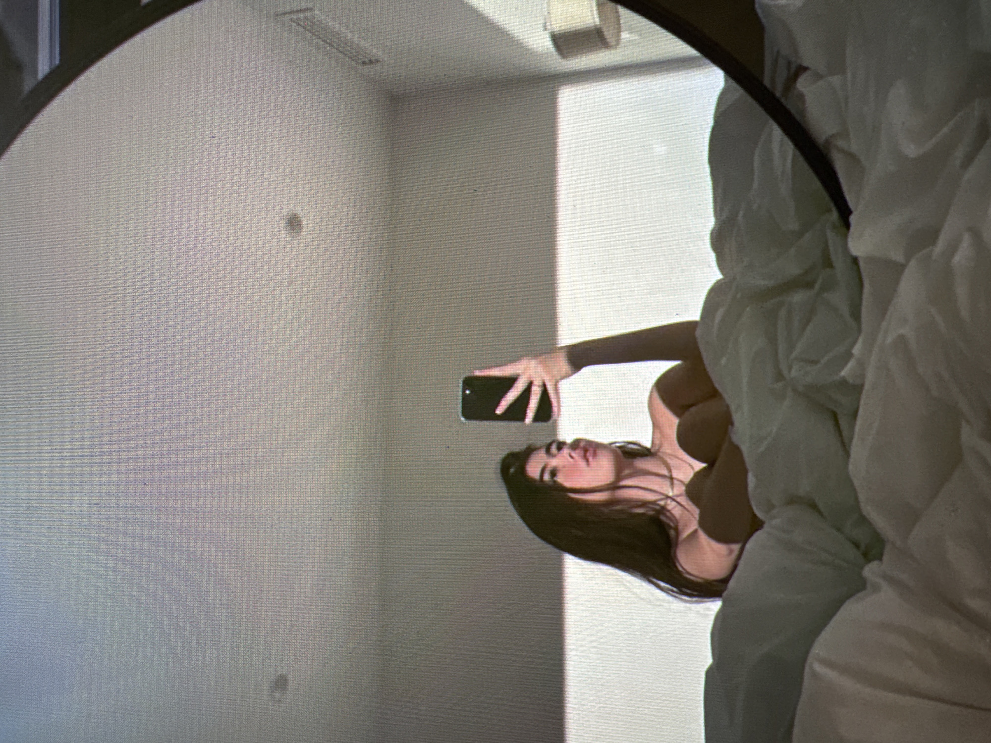 The image shows a young woman with long dark hair lying in bed and taking a selfie with her phone. She is wearing a light-colored top and a necklace. The room is lit by natural light, creating a soft glow. The bed is covered with white linens. The image is taken from a low angle, looking up at the woman. The location in the city of Castelo Branco, Portugal is not specified in the image.