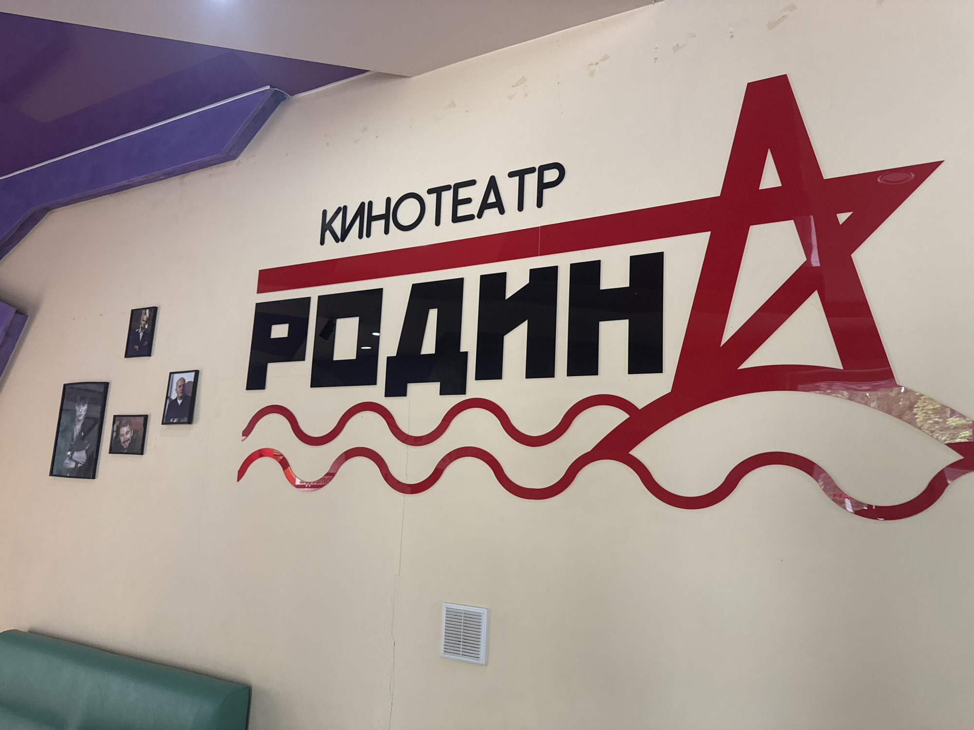 The image shows a wall in what appears to be a public building, specifically a cinema or theater. The wall is painted a light beige color. On the wall, there is signage in Russian, which reads "KinoTEATP" (Cinema/Theater) above the word "Родин" (Rodin). The word "Родин" is in bold, black letters, and below it is a decorative wavy red design. To the right of the text, there is a large red star design. To the left of the signage, there are three framed portraits hanging on the wall. Below the portraits, a green-colored seating area is visible. Below the wavy design is a white air vent.
The image is likely taken in Birobidzhan, Russia, given the content of the Russian signage on the wall.