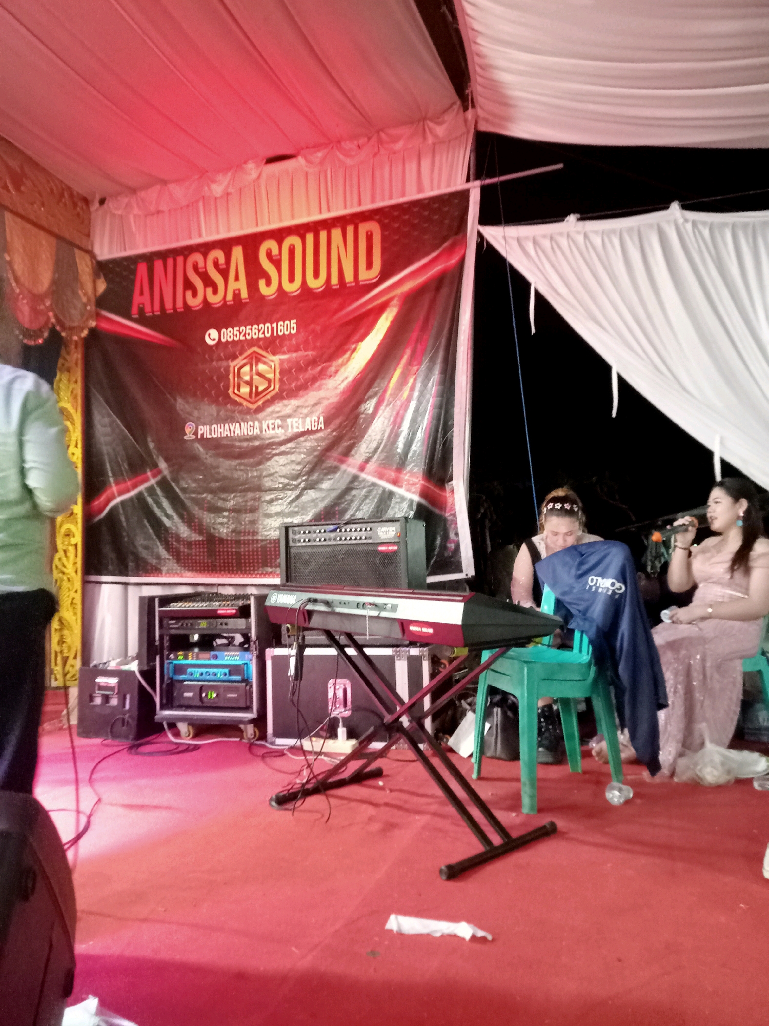 This image depicts an outdoor stage setup in Mongolato, Indonesia. A large banner with the text "ANISSA SOUND" and a phone number "085256201605" is prominently displayed. Below the banner, audio equipment, including a keyboard and amplifiers, is set up on a red carpeted stage. Two women are seated on chairs. One woman, in a pink dress, is holding a microphone and appears to be singing or speaking. The other woman, wearing a black top, is looking down. The stage is illuminated by red lighting from above, and white drapes hang on the right side. The background is dark, suggesting it is nighttime. The overall scene appears to be an event or performance.