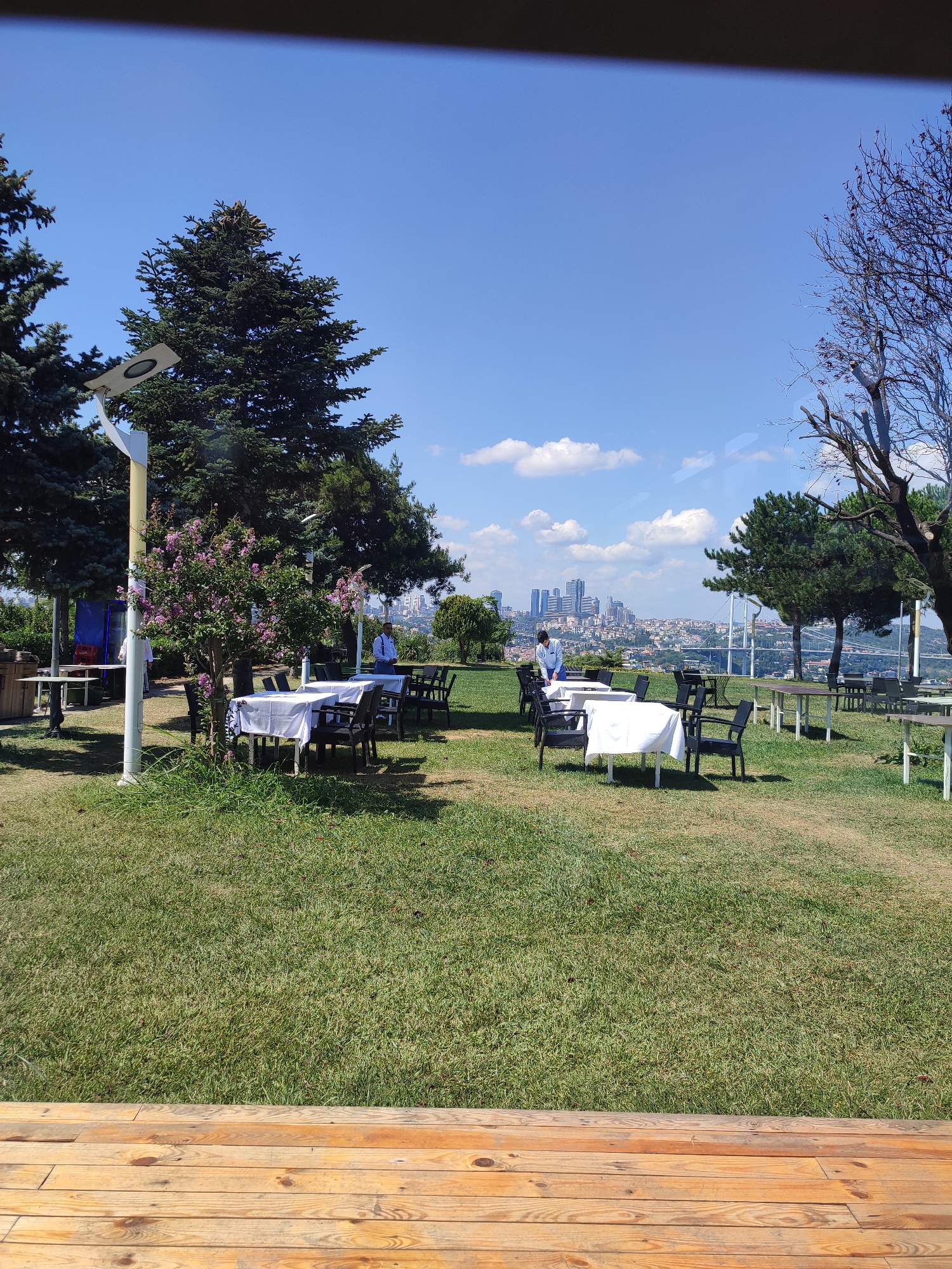 The image depicts an outdoor seating area with tables and chairs arranged on a grassy lawn, overlooking a cityscape and a body of water. The tables are covered with white tablecloths, and some are set with chairs. In the background, a dense urban landscape with high-rise buildings and a bridge is visible under a clear blue sky with scattered clouds. Several individuals are present, appearing to be staff setting up or attending to the tables. The foreground shows a wooden deck. The location is identified as being in Üsküdar, Turkey, suggesting the view is of the Bosphorus Strait and the European side of Istanbul. The overall scene conveys a peaceful, pleasant atmosphere for dining or recreation in an urban park setting.
