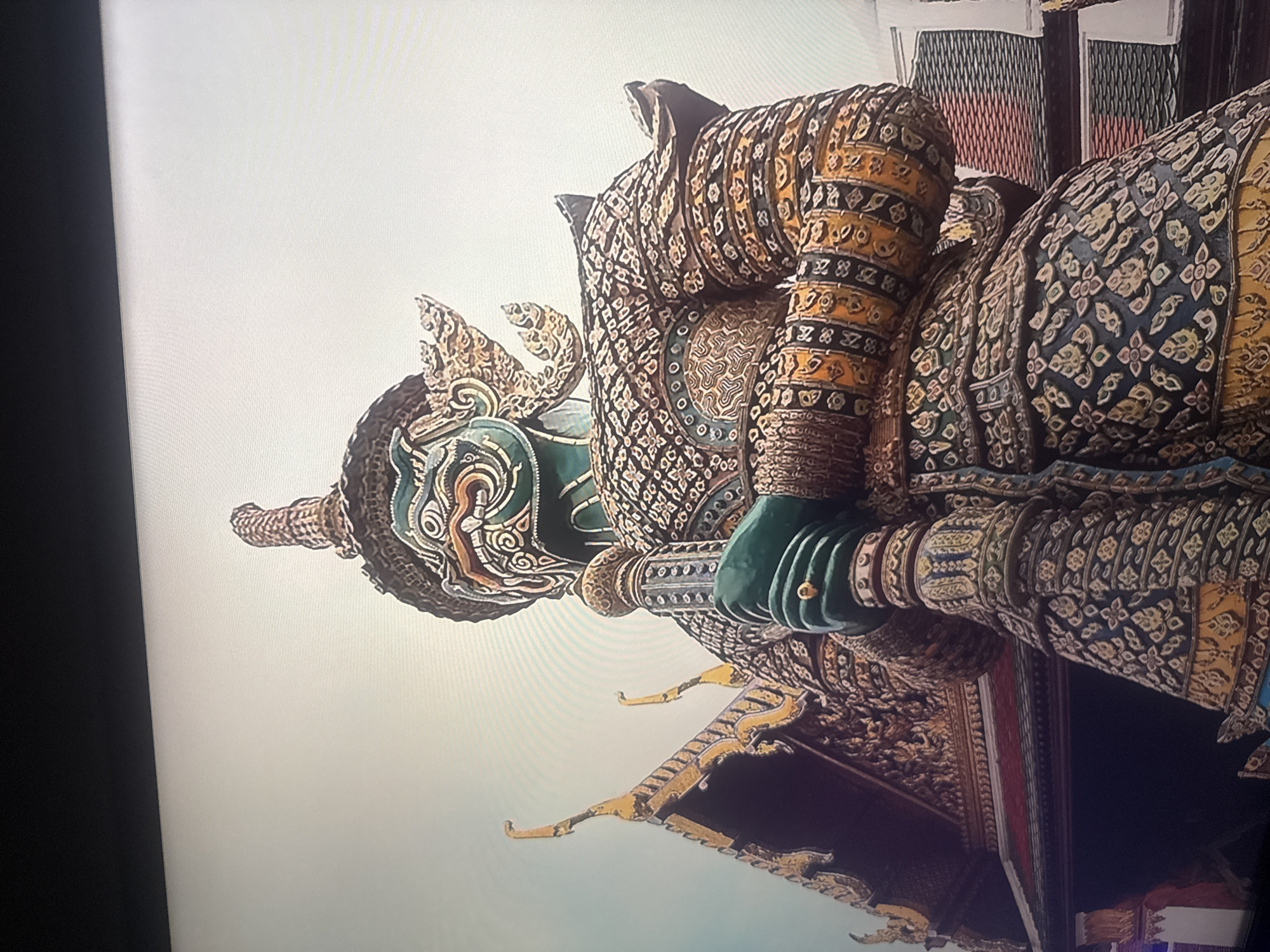 The image shows a close-up, low-angle shot of a large, ornate statue of a mythical warrior, possibly a yaksha from Thai mythology, holding a staff. The statue is elaborately decorated with intricate patterns and vibrant colors, featuring a green face with exaggerated features and a golden headdress. Behind the statue, a portion of a traditional Thai temple roof with gilded carvings is visible. The statue and temple architecture suggest a religious or cultural site, likely in Thailand. There is no indication of the city of Herzliya, Israel, in this image. The background is a pale, hazy sky, and the foreground shows a dark, blurry area, possibly a screen or frame.