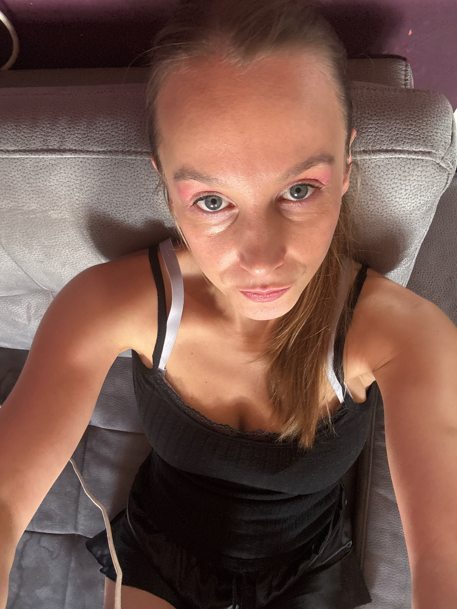 A woman with blonde hair tied in a ponytail and light blue eyes is looking at the camera. She is wearing a black tank top with lace trim and white and grey straps. Her lips are painted with pink lipstick. She has pink eyeshadow on her eyelids. The woman is sitting on a grey couch with a textured surface. The background is dark, with a hint of purple at the top. There is a white cable running down the left side of the image. The overall scene appears to be an indoor setting. There is no information to suggest the location is in Überherrn, Germany.
