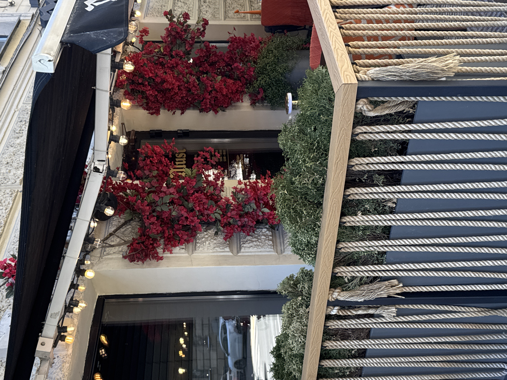 The image shows a close-up view of an awning over a storefront, adorned with strings of lights and artificial red flowers. The awning is dark blue and made of fabric, with a metal frame. Below the awning, a window reveals the interior of the establishment, with some of the lights reflecting in the glass. The storefront appears to be decorated with more red flowers and greenery, suggesting an outdoor or semi-outdoor seating area. To the right of the storefront, a decorative wooden structure with thick ropes stretched across it is visible. The ropes are beige and appear to be made of natural fibers, creating a textured, nautical-inspired element. The lighting in the image suggests it might be evening or a dimly lit day, as the string lights are illuminated. The overall scene evokes a cozy and inviting atmosphere, perhaps a cafe, restaurant, or shop.
There is visible text on the awning, though it is partially obscured and difficult to read clearly. It appears to be a logo or name, possibly in white lettering. The text "Just" is also visible in the reflection of the window.