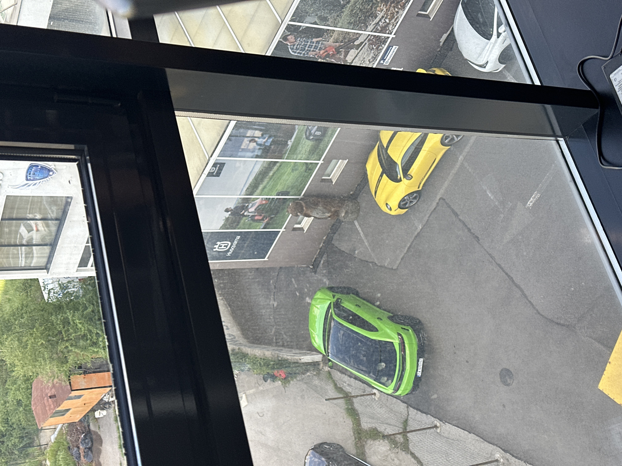 The image, apparently taken through a window of a building, shows an external scene of a street or parking area. Several vehicles are visible in the scene. A green car is parked in the lower part of the frame. A yellow car is located in the upper portion of the frame, near what appears to be a poster or advertisement showing images of Husqvarna products and people using gardening equipment, suggesting this image could be of Husqvarna products. The location of the image could potentially be inferred from the license plate seen on the green car, reading "CA 0707GT", and additional information from the surrounding buildings. Given the identification of a Bulgarian license plate, the location is Sofia, Bulgaria. The weather conditions cannot be clearly determined from this image, although the surface appears to be dry. There is no visible human activity, although there are people depicted in the advertisement.