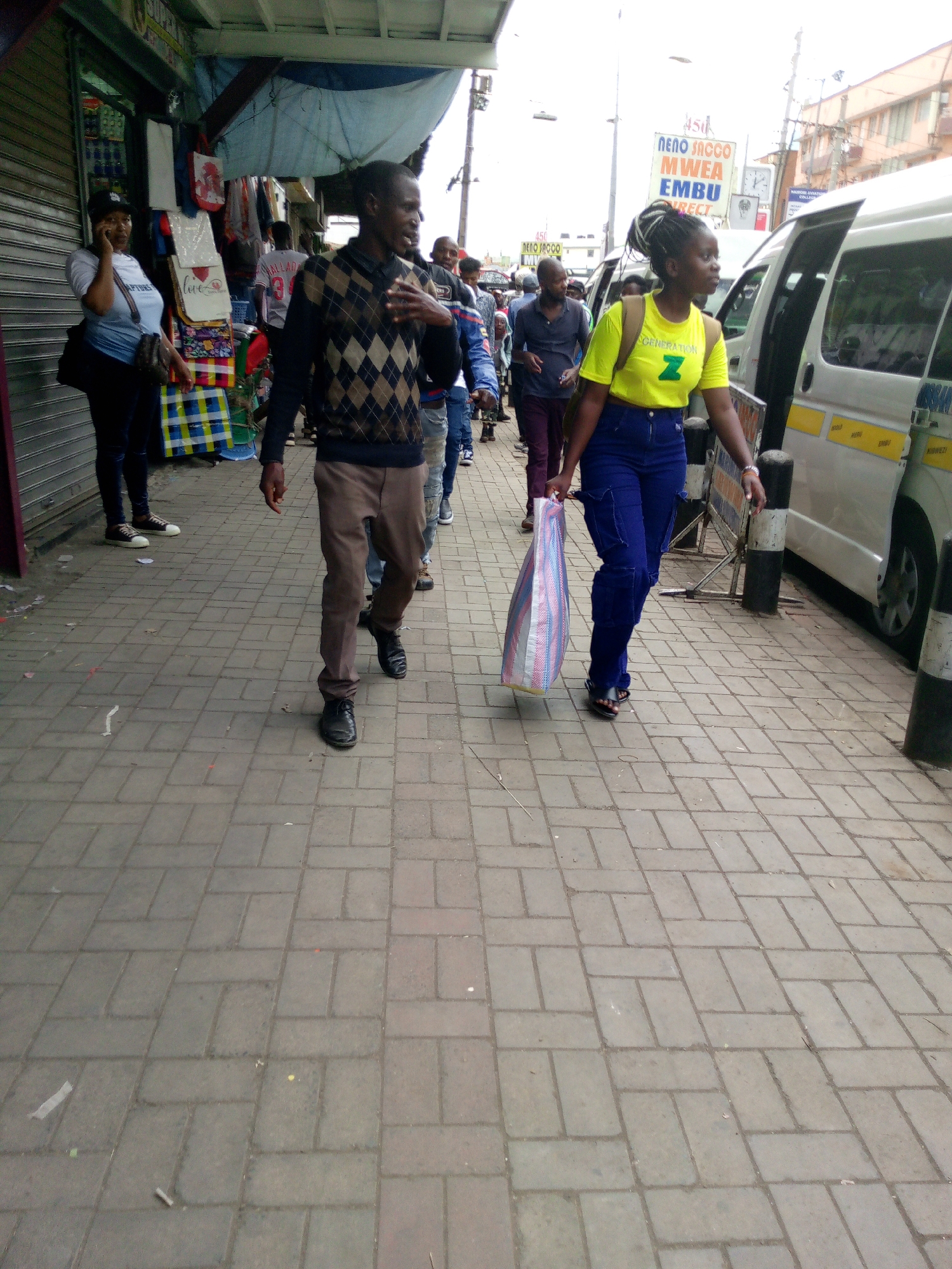 The image captures a street scene in Nairobi, Kenya. A man in a patterned sweater and brown pants walks beside a woman wearing a yellow shirt and blue cargo pants, who carries a striped bag. They are walking on a sidewalk paved with gray rectangular bricks, next to a street with parked white vans. Other pedestrians can be seen in the background.
On the left, a storefront with a partially rolled-down shutter displays merchandise, including clothing and bags. Above the storefront is a blue awning. A woman wearing a black baseball cap and a blue t-shirt with the word "Raptors" is talking on her phone.
In the background, a sign reads "NENO SACCO MWEA EMBU." The sky is overcast. The scene conveys a sense of everyday life in an urban Kenyan setting.