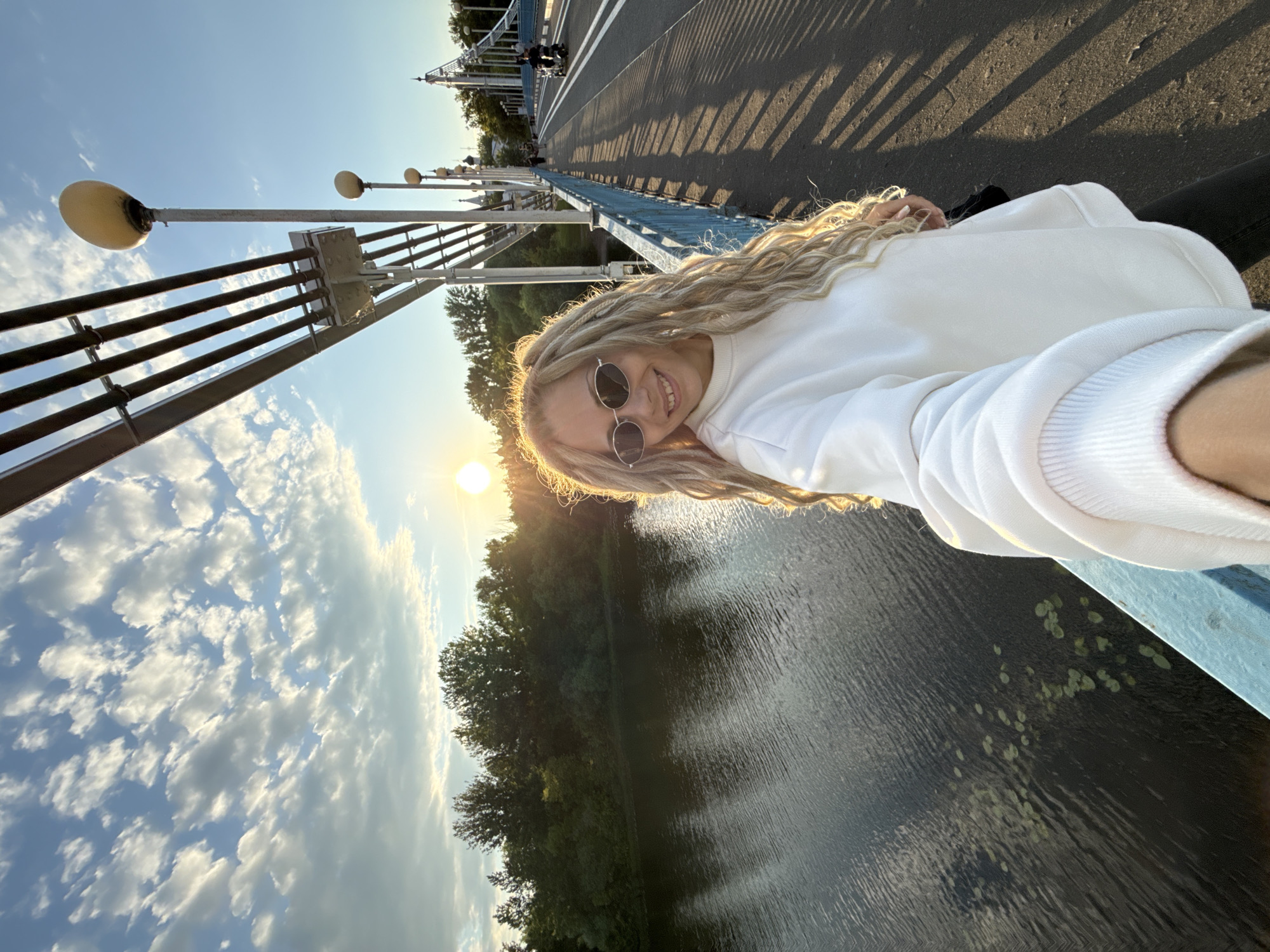 A sun-drenched selfie captures a young woman smiling on a bridge, set against a backdrop of a river and a clear, partly cloudy sky. The woman, with long, light-colored curly hair or dreadlocks, wears round sunglasses and a white sweatshirt. She is positioned close to the camera, with the bright, low-angle sun behind her, creating a warm glow and some lens flare.
The perspective is tilted, showcasing the bridge's structure. Blue railings are visible closest to the viewer, with lampposts featuring round, globe-like lights extending along the bridge. Further along the bridge, long shadows stretch across the asphalt road, indicating the late afternoon or early evening time, often referred to as the "golden hour." Distant parts of the bridge, possibly including a decorative tower, can be seen.
Below the bridge, a body of water, likely a river or lake, shimmers with reflections of the sky and light. Ripples are visible on the water's surface, and patches of green aquatic plants float near the bridge's edge. The far bank is densely forested with green trees under a bright blue sky dotted with scattered white clouds. The overall mood is pleasant and joyful, capturing an outdoor moment. No visible text is present in the image. The location context provided is Шебунино, Russia.