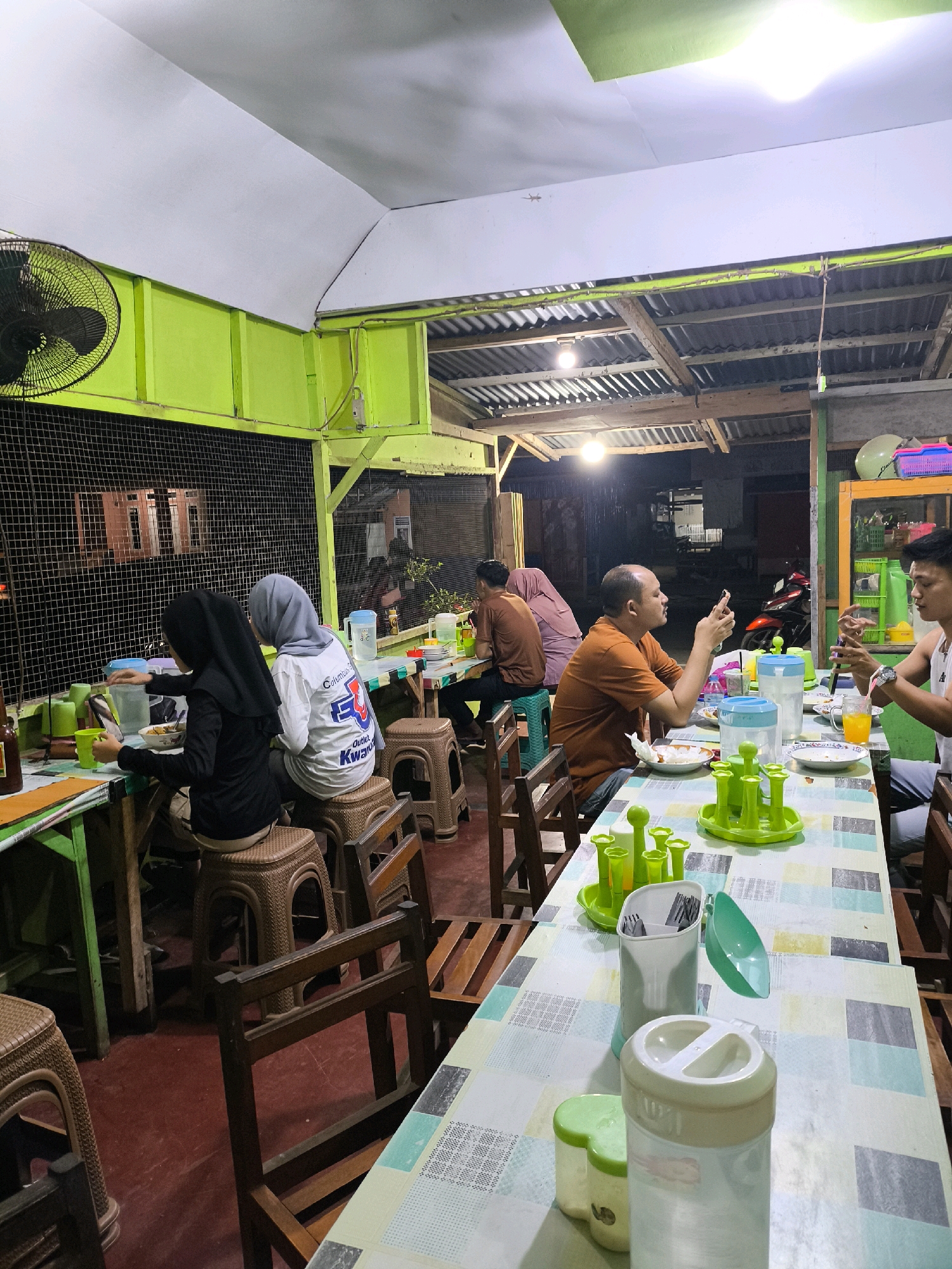 The image depicts an outdoor dining area at what appears to be a casual eatery in Moluo, Indonesia. The setting is dimly lit with overhead lighting and natural light filtering in from the right. Several tables are arranged, covered with checkered tablecloths. People are seated at the tables, engaged in dining and conversation.
On the left side of the image, two individuals are seated at a table. One person, wearing a black hijab, is looking down at their phone and holding a spoon. The other person, wearing a grey hijab and a white t-shirt, is also looking towards the table. Green plastic cups and bottles of water are visible on this table.
Further into the scene, more tables and patrons are present. A man in an orange t-shirt is seated at a table, holding his phone and looking at it. Another man, wearing a white tank top, is seated opposite him, gesturing with his hands. This table has plates of food, glasses of orange juice, and various containers. A red motorcycle is parked in the background.
The environment features a corrugated metal roof supported by wooden beams. The walls are painted a bright green, and a large fan is mounted on the left side. The floor is a reddish-brown color. The overall atmosphere suggests a bustling, local dining experience.