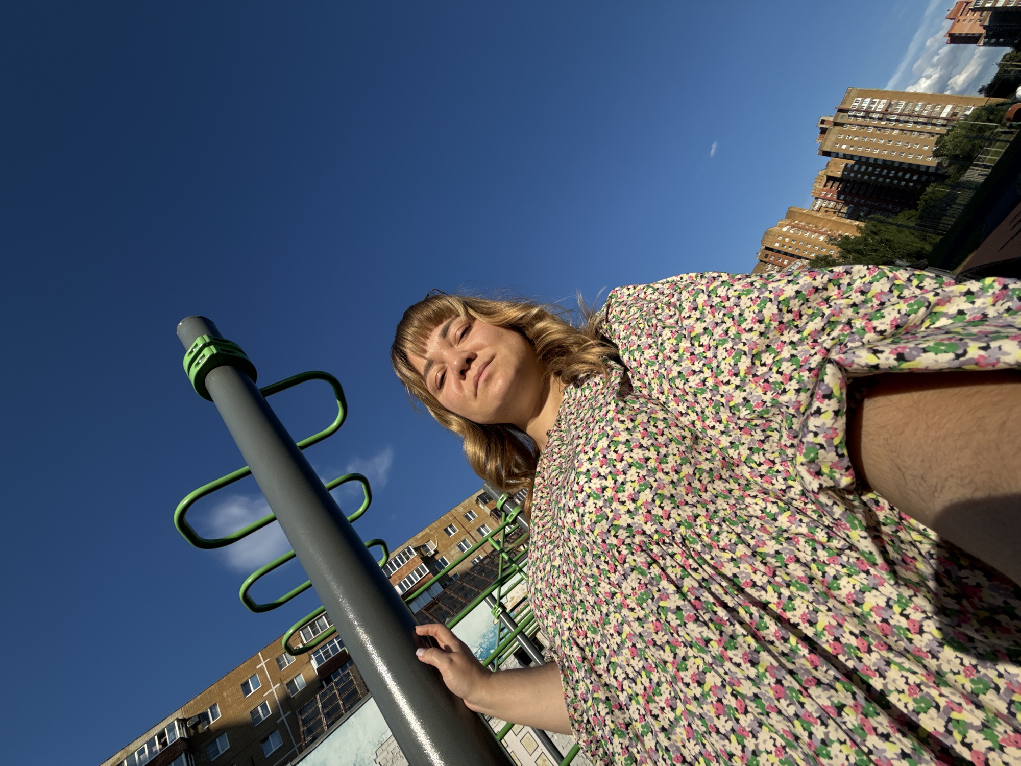 A woman with blonde hair and bangs, wearing a floral dress, stands outdoors in front of a bright blue sky. She is holding onto a gray horizontal bar with green attachments, part of an outdoor fitness equipment setup. In the background, tall apartment buildings are visible, suggesting an urban or suburban environment. The perspective is from a low angle, looking up at the woman and the sky.