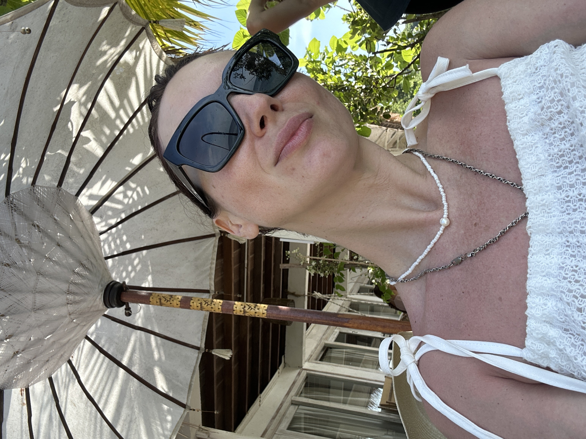 A fair-skinned woman with dark hair and wearing dark sunglasses is seen from a low angle, tilted, against a background featuring a white umbrella, vegetation, and a building. She wears a white lace top with tied shoulder straps and two necklaces. The umbrella is white and patterned with dark lines. Green foliage and a portion of a hand are visible in the upper portion of the image. In the background behind the umbrella, part of a white building with windows is visible. This scene is likely in Kuta Selatan, Indonesia, given the type of umbrella, vegetation, and architecture visible.